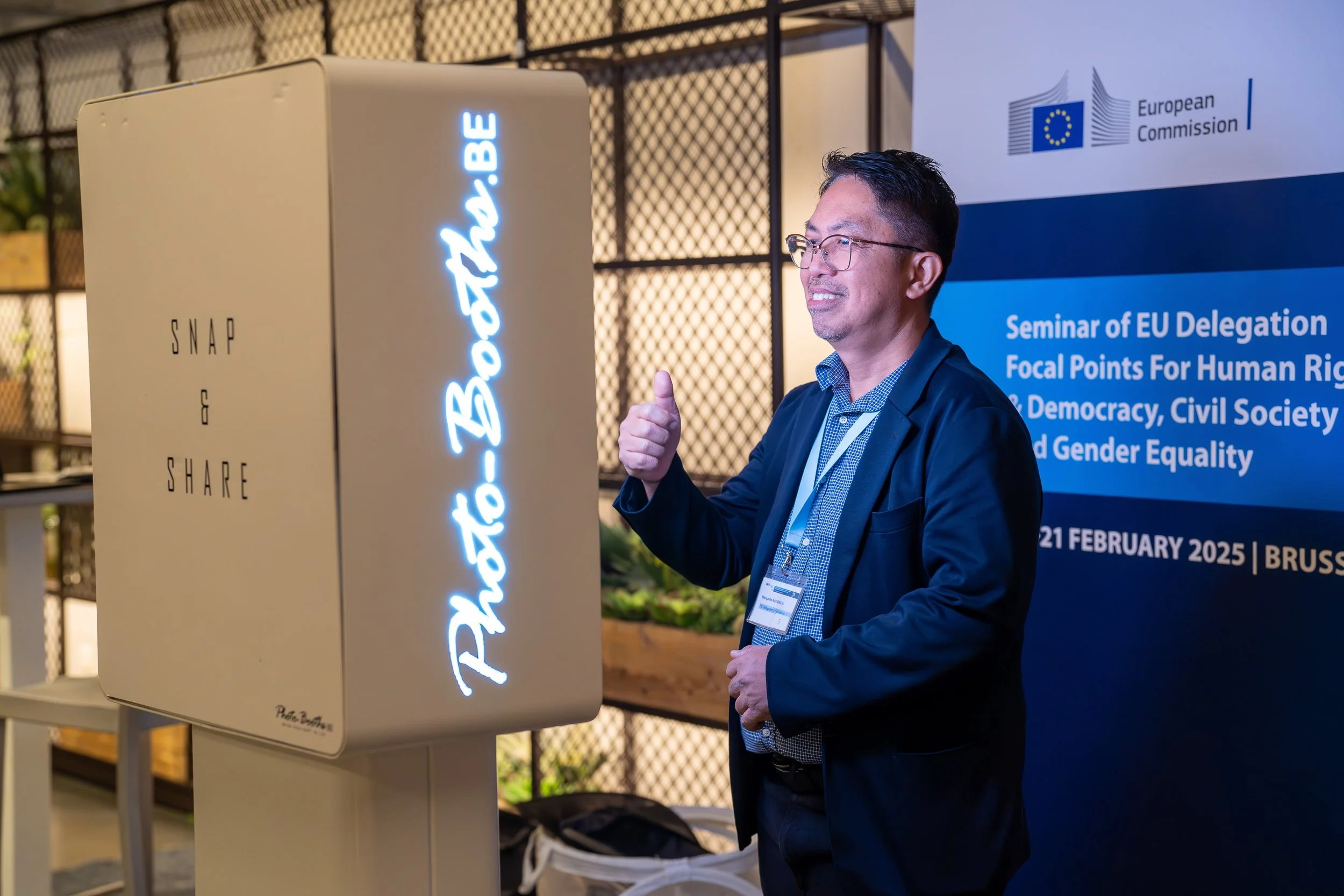 A man in glasses and a dark suit giving a thumbs-up while standing next to a poster and a white box with illuminated text at a European Union seminar in Brussels. EU Delegation Seminar on Human Rights 2025, Democracy & Gender Equality Brussels