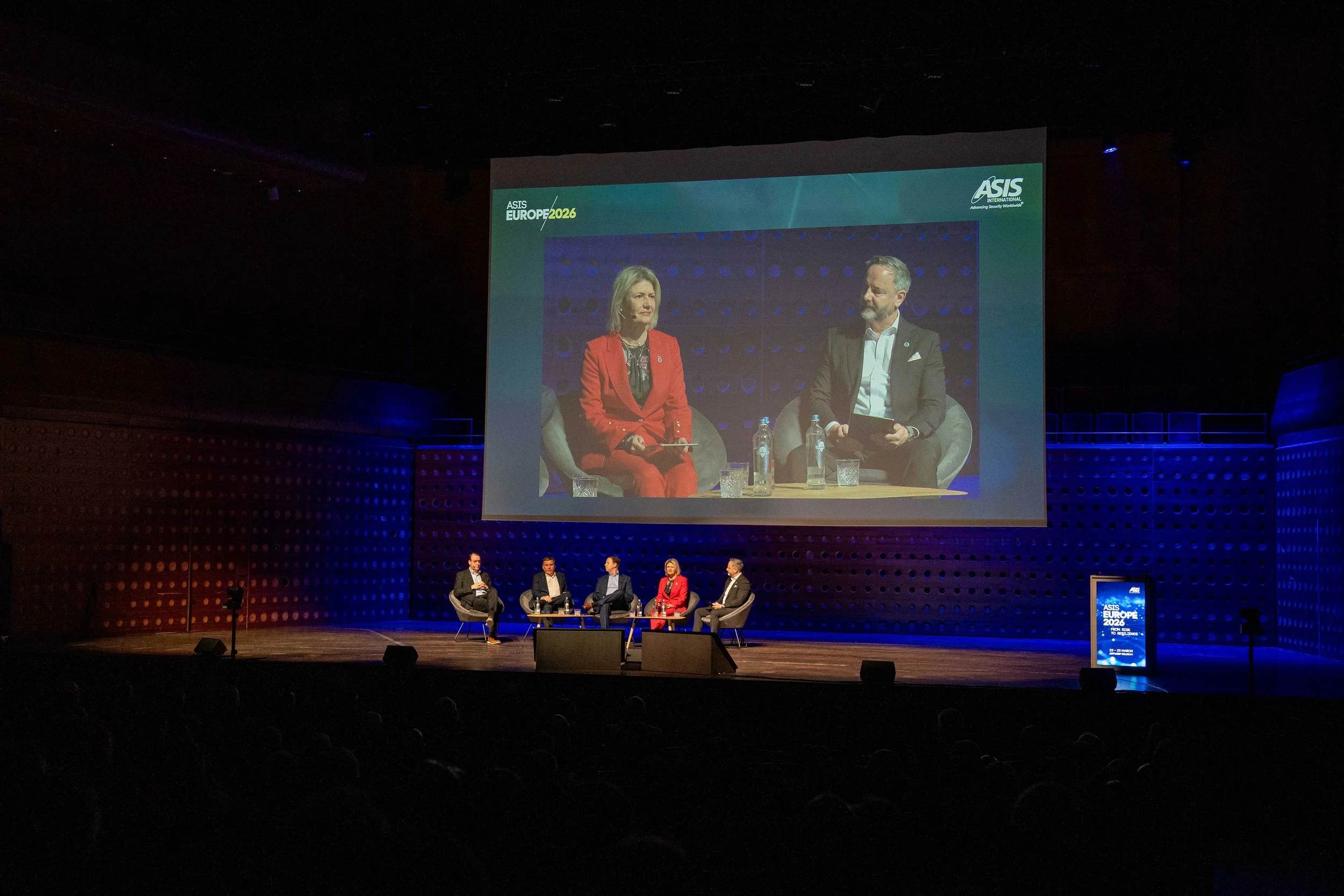 A large conference hall with a panel discussion taking place on stage. Five people are seated on chairs, and a woman in a red suit is speaking. There is a large screen behind them showing a live feed of the panel, with the event titled "ASIS Europe 2