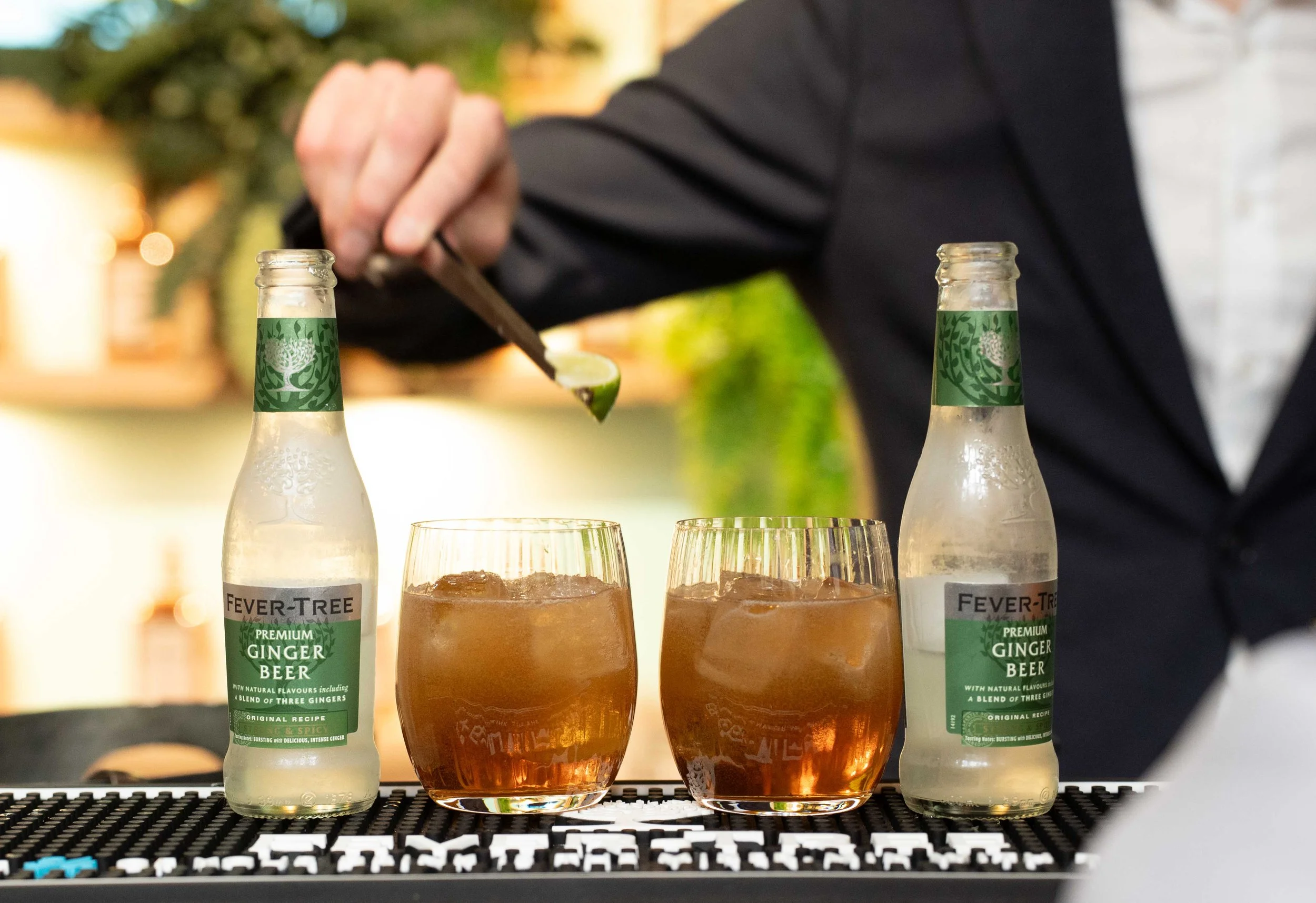 Two bottles of Fever-Tree premium ginger beer on a bar or counter, with two glasses filled with ginger beer and ice in front of them. A person in black clothing is pouring a drink from a spoon in the background. Michelin guide Ceremony 2026 Antwerp, 