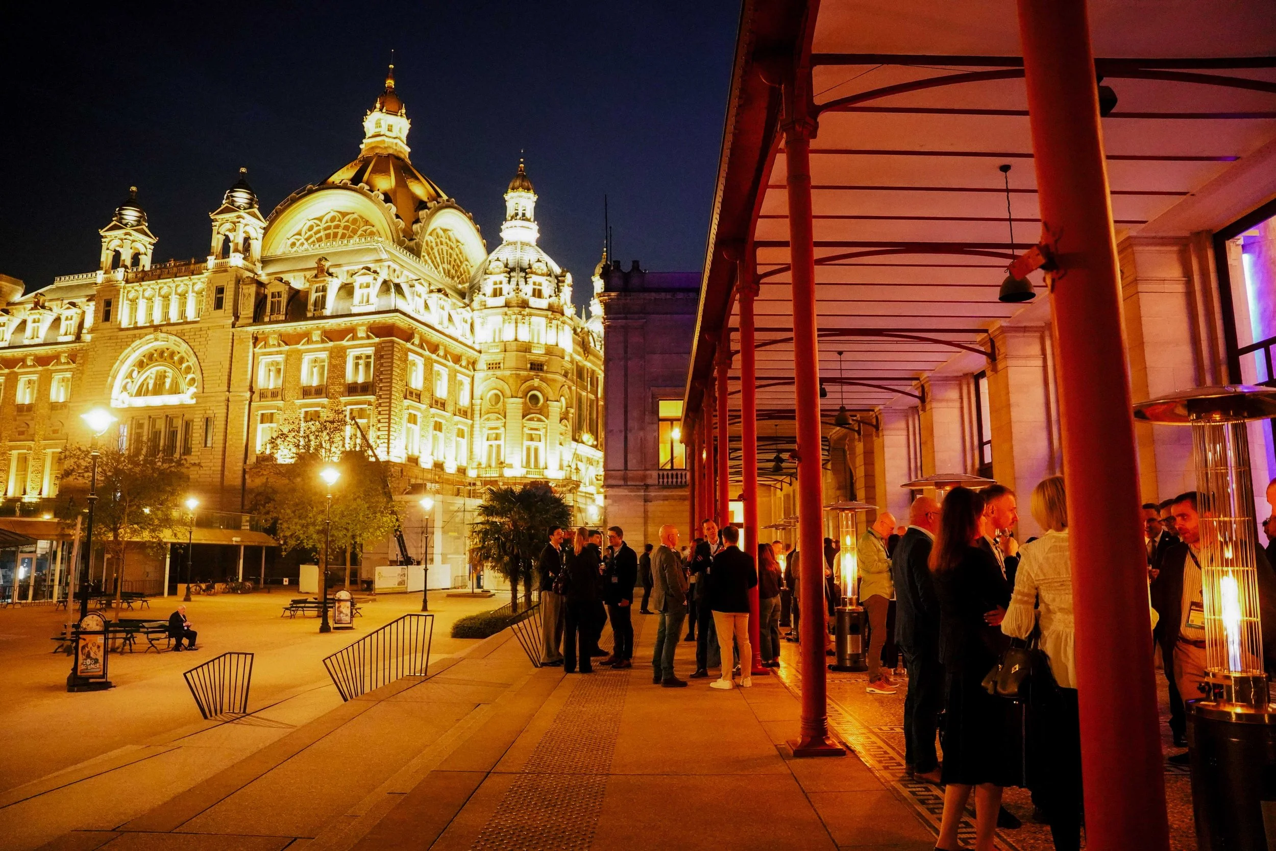 Night view of an illuminated historic building with domes and towers, with people gathered and socializing in an outdoor patio area with lighting and heaters.