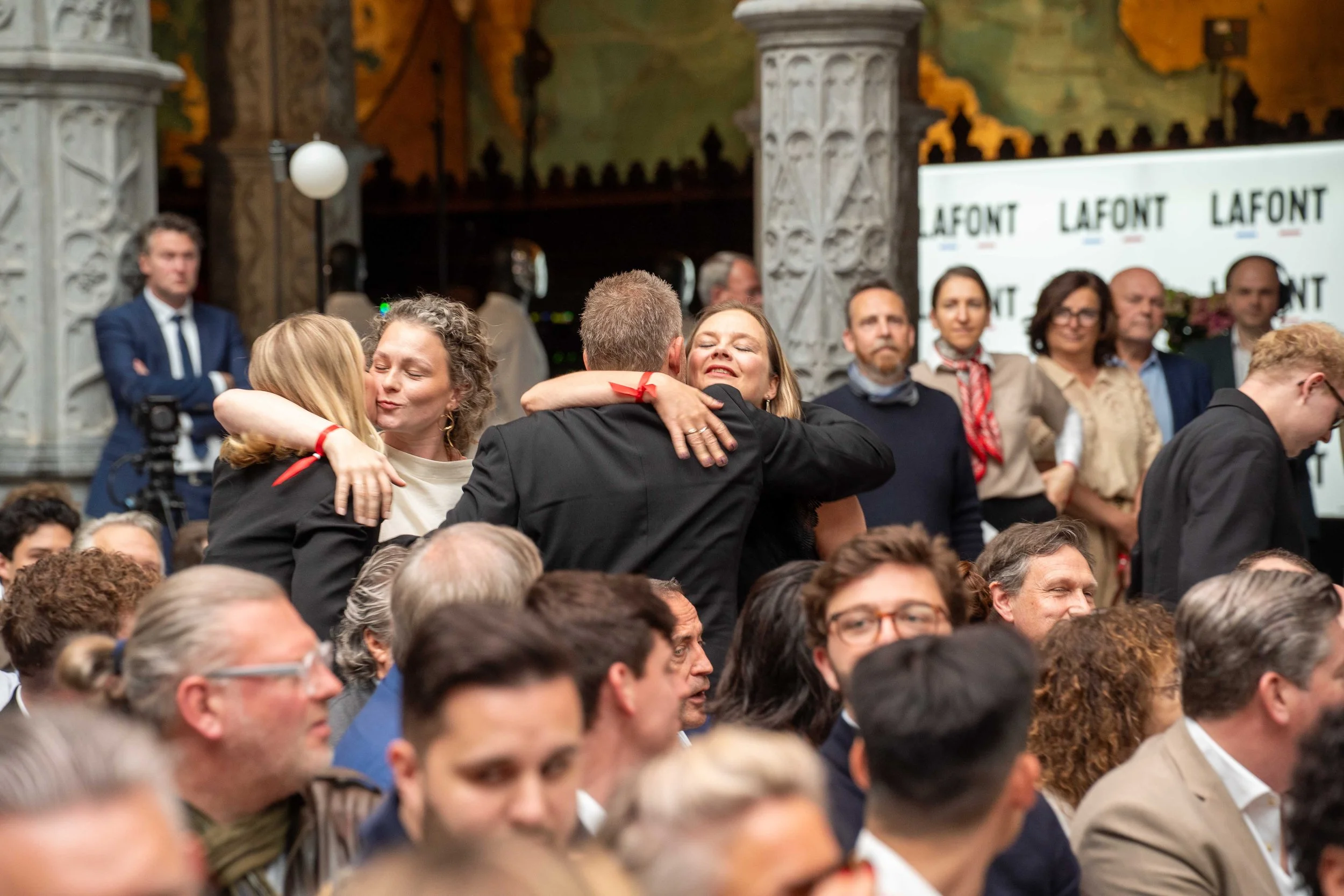 A group of people attending an event, with two women hugging in the center, surrounded by other attendees standing and watching. The background features decorative architecture and a sign that reads 'LAFONT'. Michelin guide Ceremony 2026 Antwerp, Han