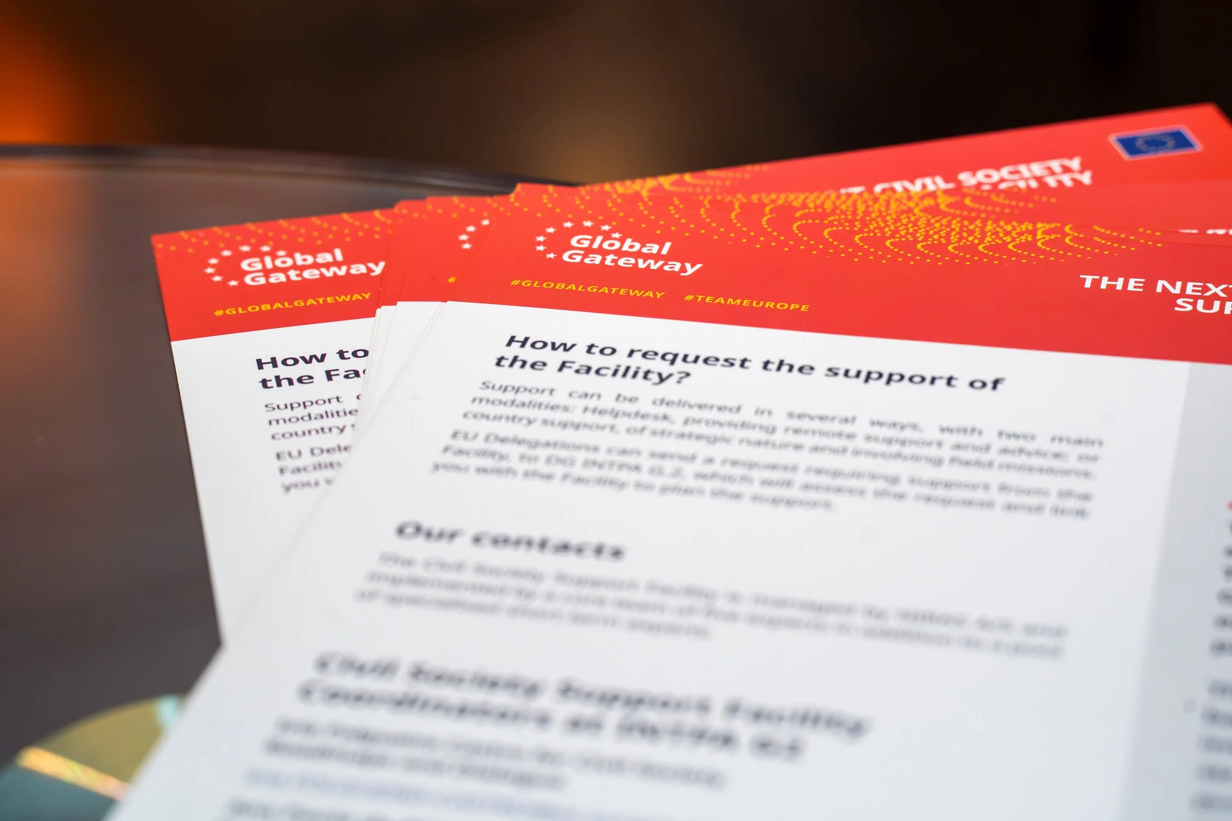 A stack of flyers or pamphlets in red and white colors promoting Global Gateway, with hashtags #GLOBALGATEWAY and #TEAMEUROPE, and text about how to request support for European facilities.
