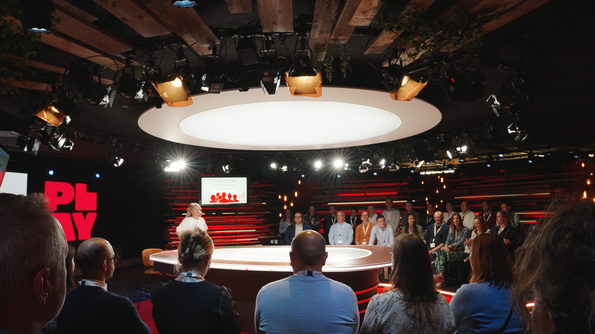 An indoor conference or panel event with a large circular stage, audience seated around, and a presenter standing on stage. Overhead lighting and screens are visible, with the audience and participants attentively listening.johnson & Johnson Finance