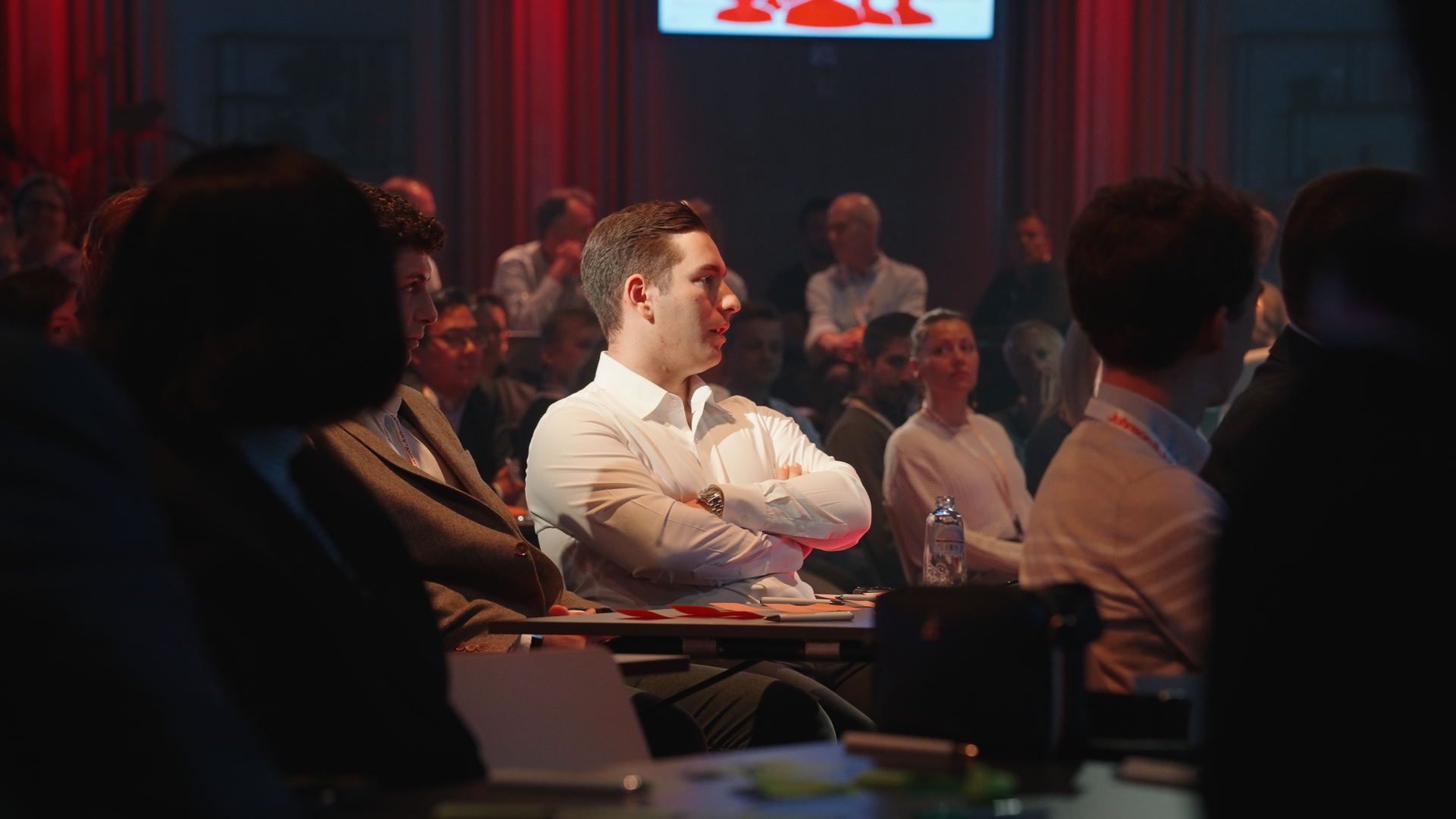 A man in a white shirt with crossed arms sitting attentively at a conference, surrounded by other audience members in a dimly lit room with a red-lit backdrop and a screen displaying a logo. johnson & Johnoson Finance day 2026 at Tv studio Play Antwe