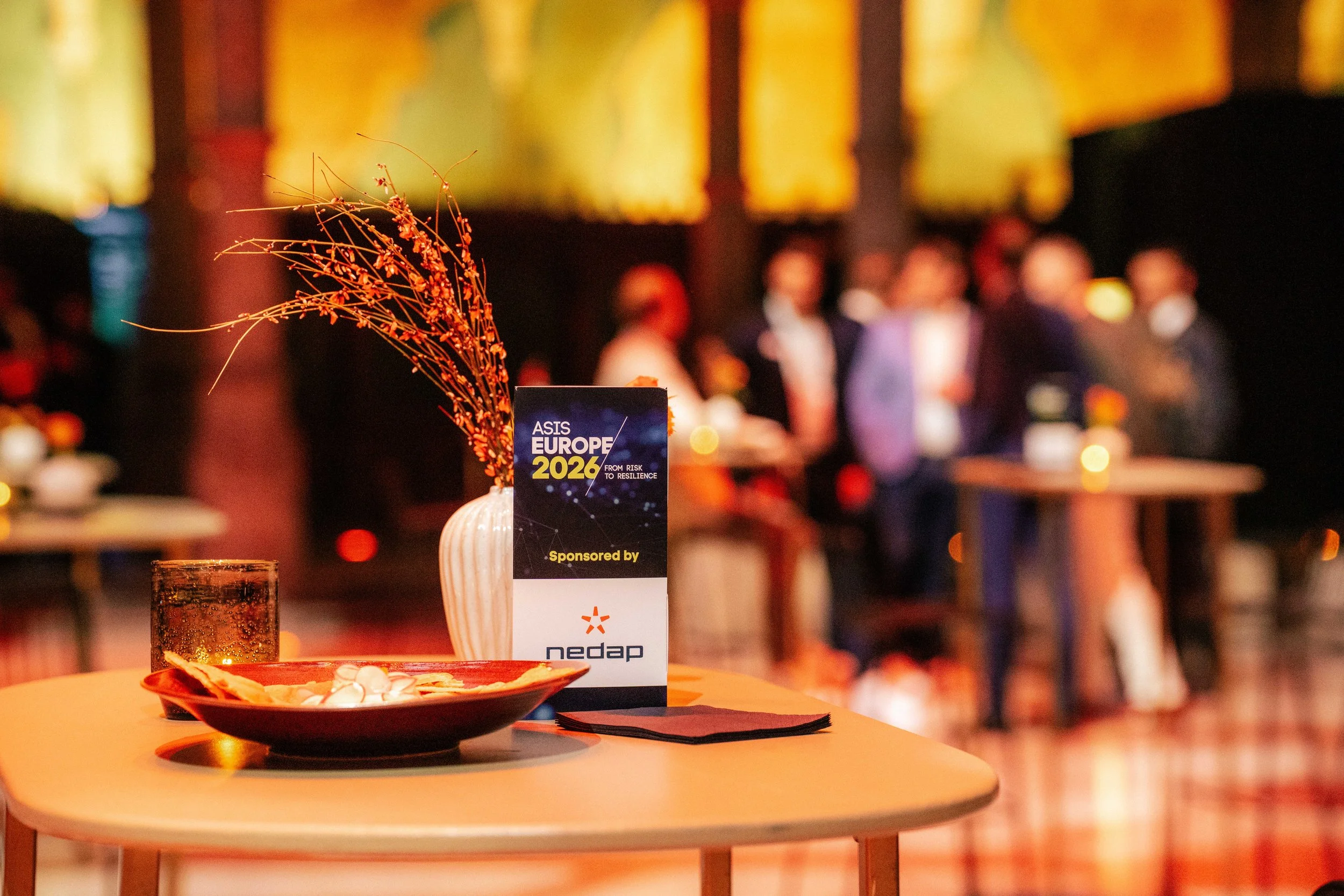 A table with a sign reading 'ASIS Europe 2026', a tall glass of a dark drink, a white vase with dried branches, and a plate of snacks, with a blurred group of people in the background at an event.