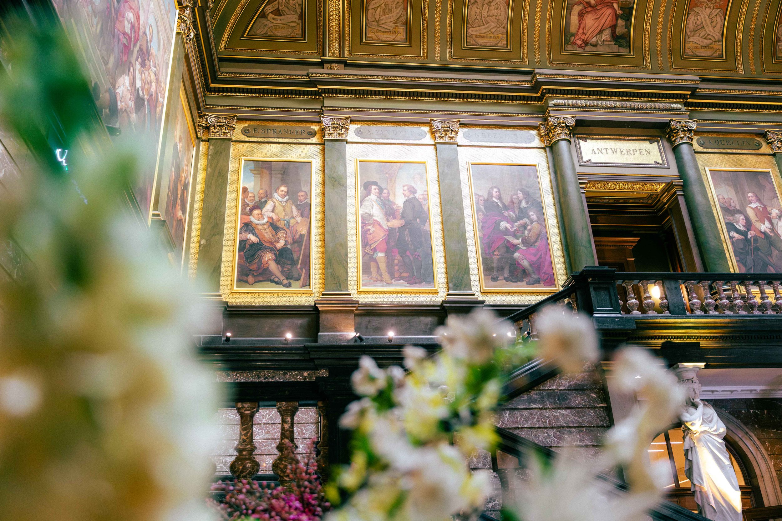 Interior of a grand, ornate building with gold-trimmed paintings and sculptures, a staircase, and floral decorations in the foreground. Network event at KMSKA for company Nelissen, Bulex, Forbo and SOPREMA.