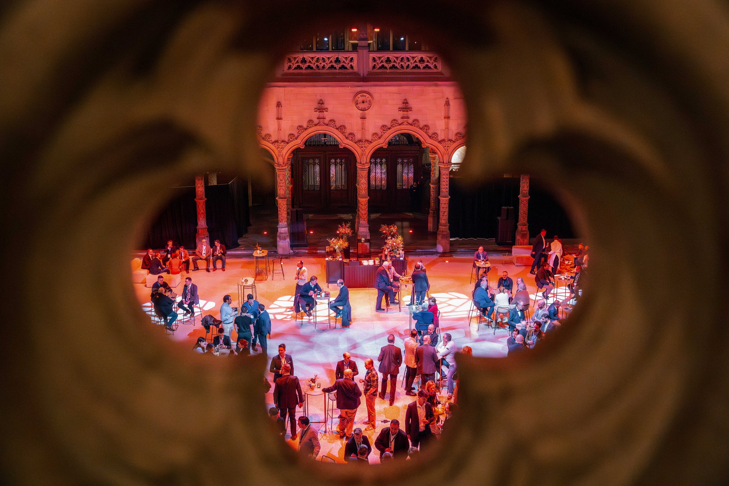 A view of an indoor social event taken through a small peephole, showing people sitting and standing in a decorated venue with ornate architecture, warm lighting, and floral arrangements.