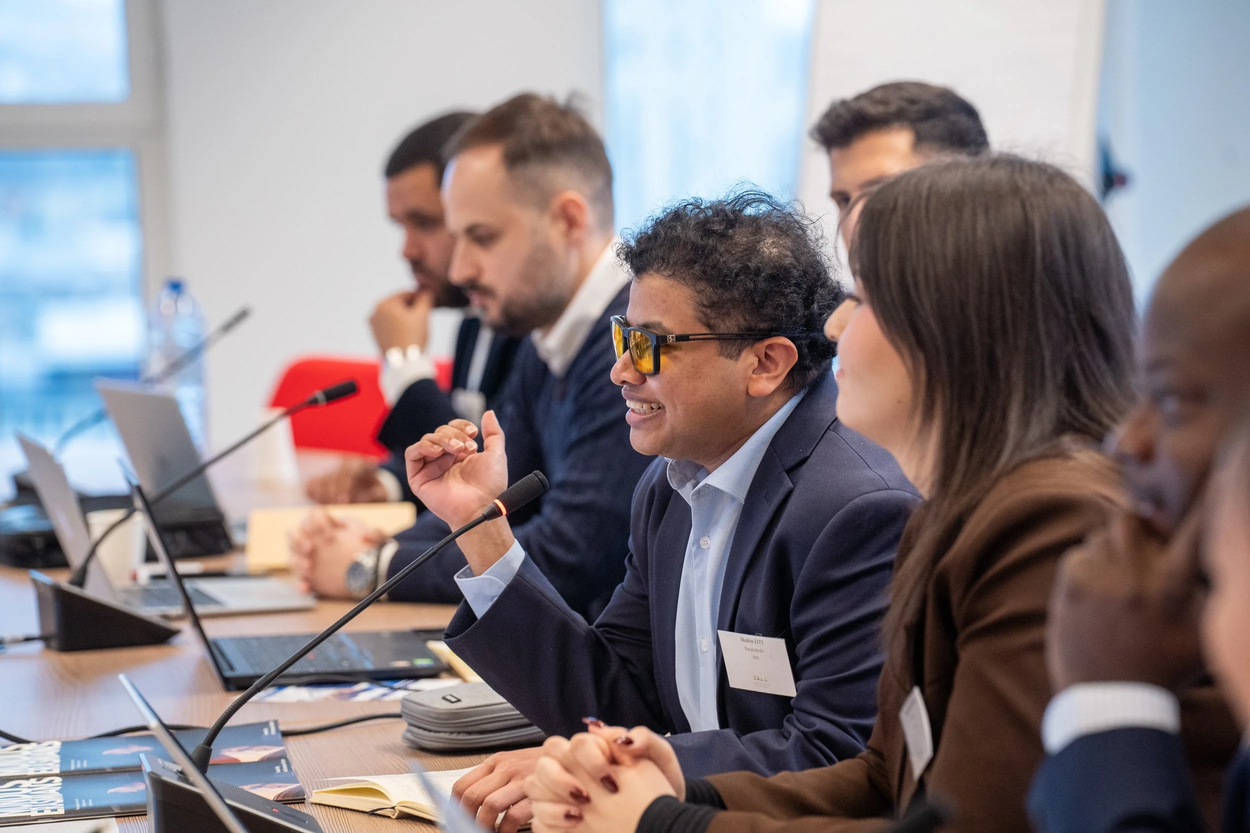 A diverse group of professionals are seated at a conference table, engaged in a meeting or discussion, with several laptops, notebooks, and microphones in front of them. Students of the Erasmus+ ESSA European Comission meeting in Brussels.