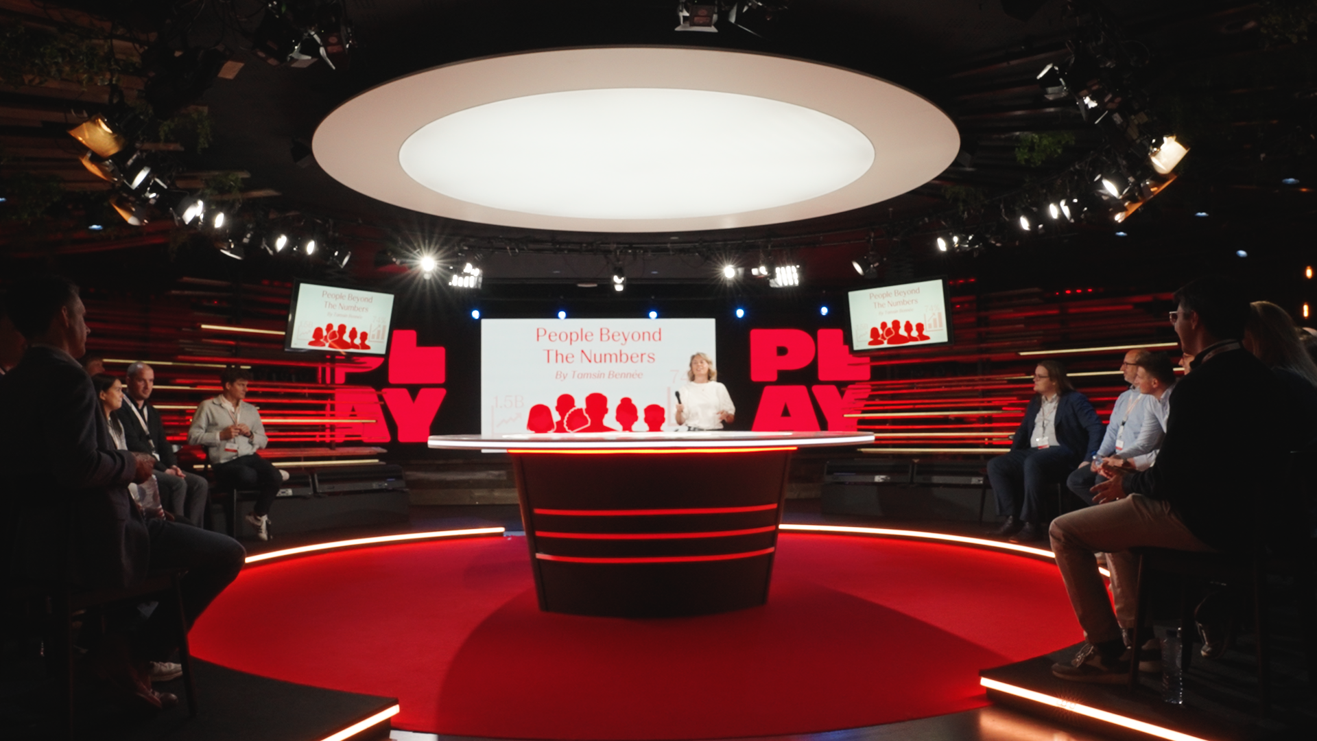 Panel discussion at a conference with a woman speaker standing behind a circular table, audience members seated around the stage, large screens displaying the presentation slide titled 'People Beyond The Numbers' by Tamsin Bennet.