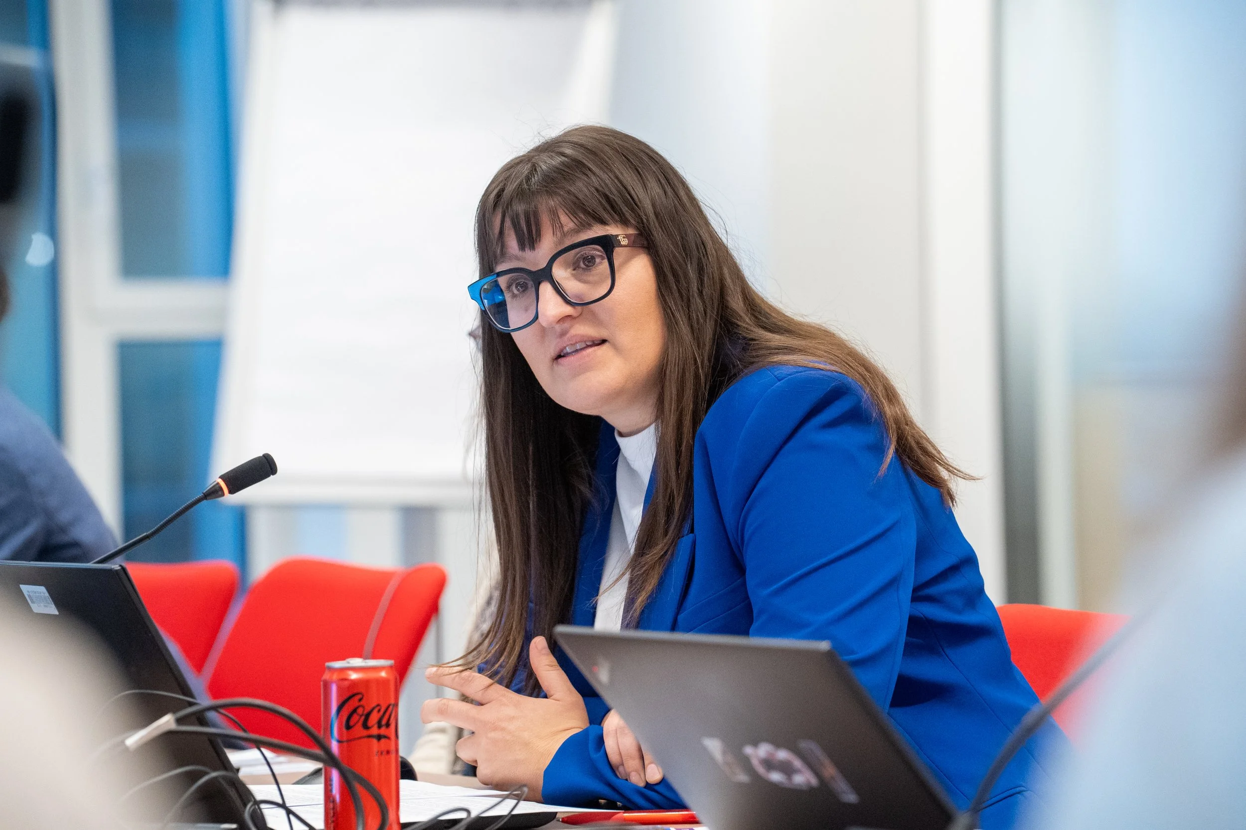 A woman with long brown hair, wearing glasses and a blue jacket, sits at a table with electronic devices, speaking into a microphone. A Coca-Cola can is on the table, and there are red chairs in the background. Erasmus+ ESSA European Comission event.