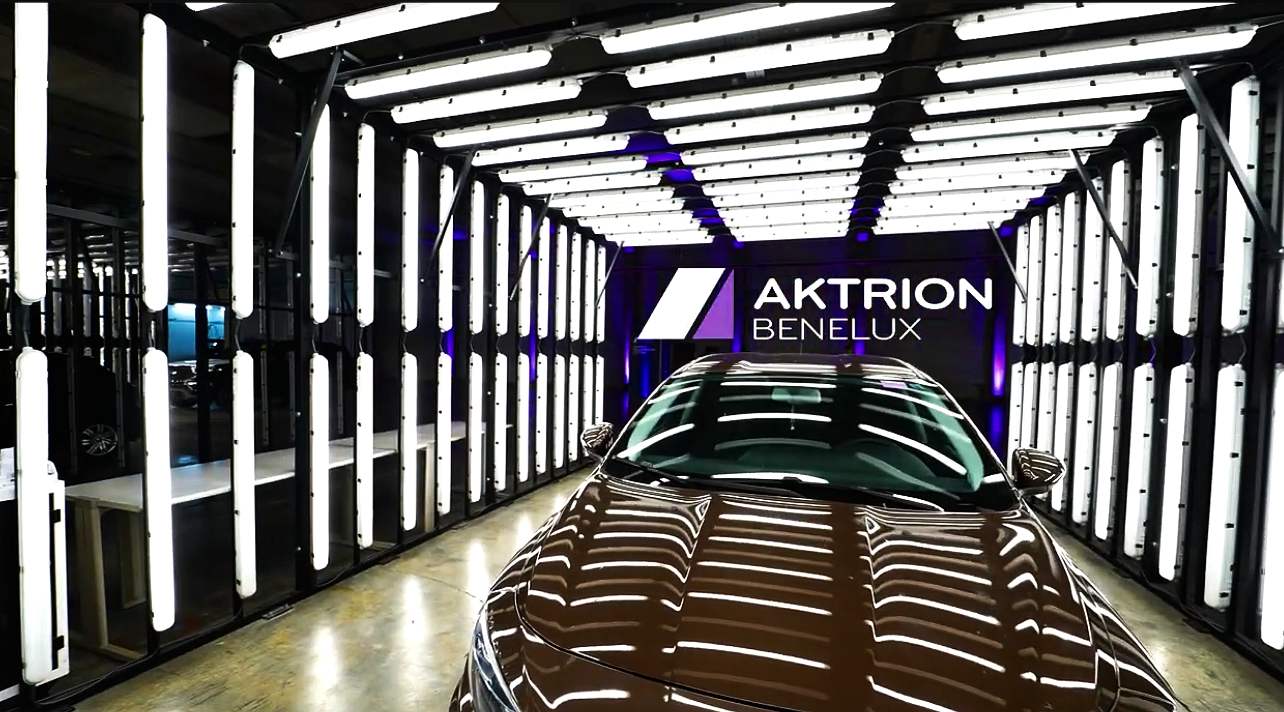 Dark-colored car parked inside a modern showroom with illuminated vertical light tubes on the walls and a sign that reads 'AKTRION BENELUX' in the background.