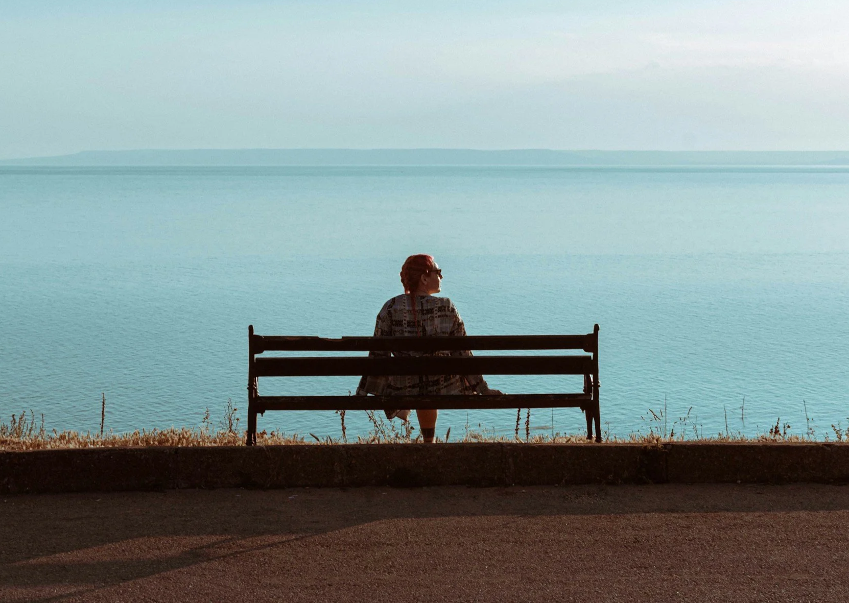 Person sitting quietly on a bench beside the water, looking out across the ocean.