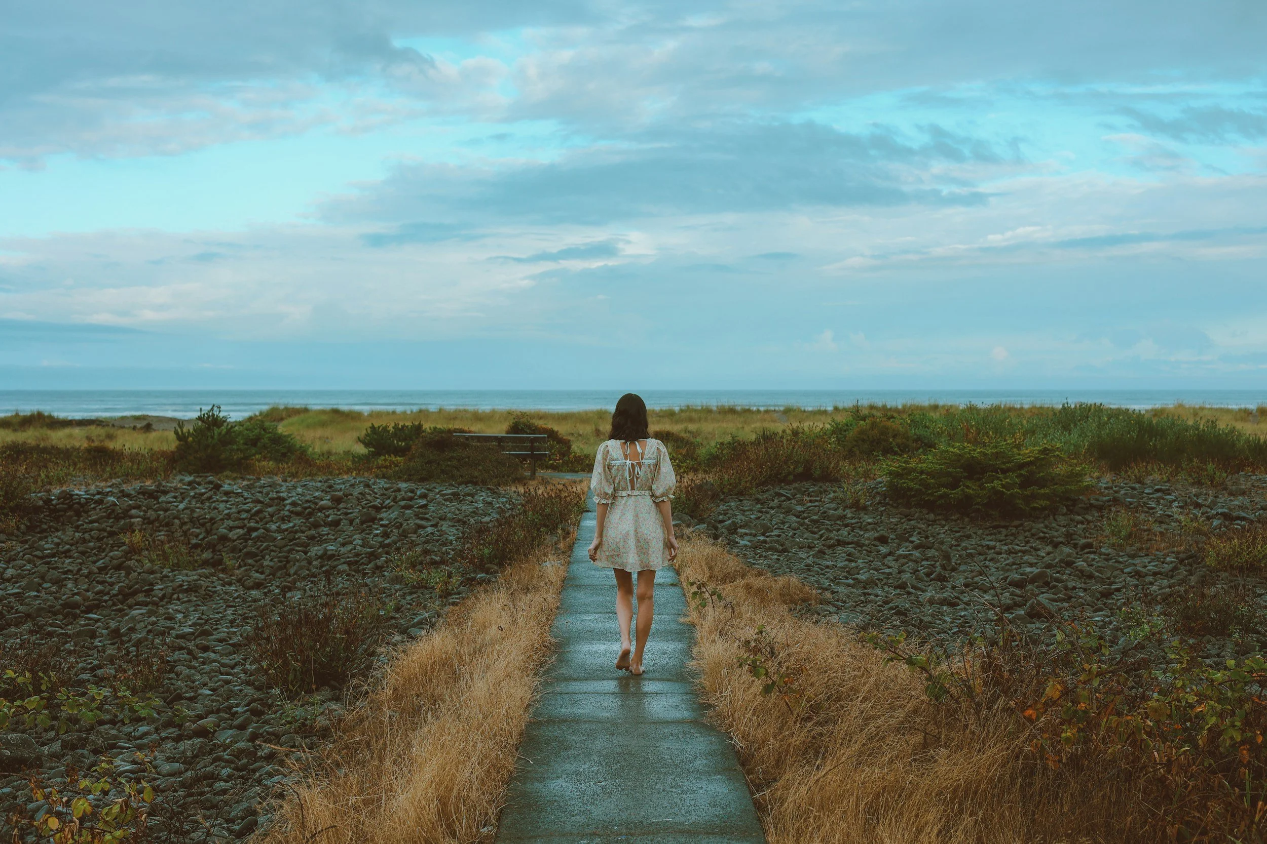 Woman walking barefoot along a stone path towards the sea.