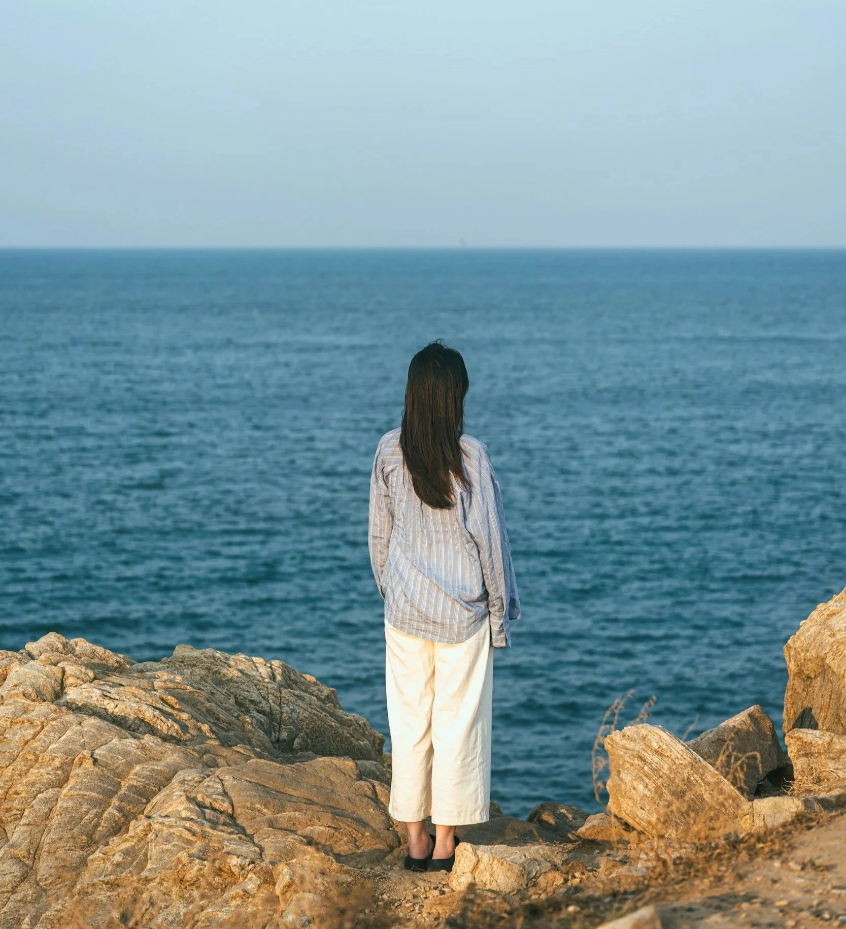 Woman standing on rocks beside the sea, looking thoughtfully across the water.