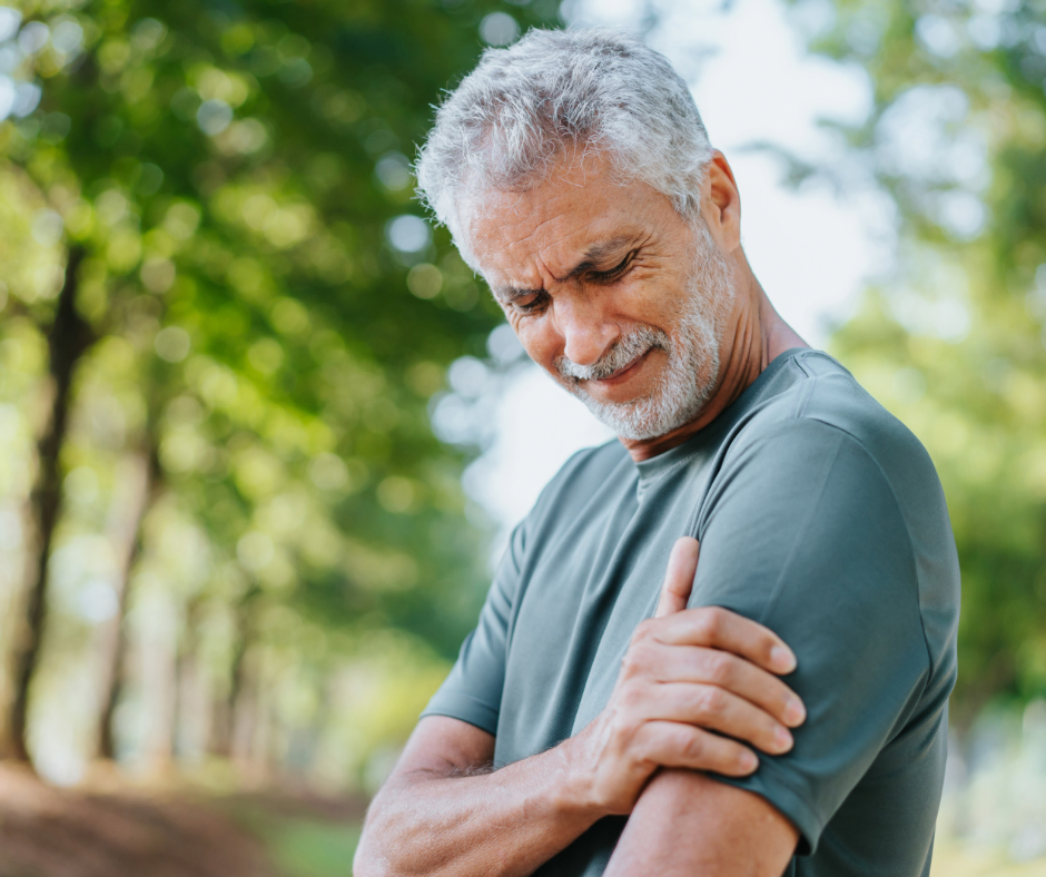 Older man outdoors, clutching his shoulder and grimacing in pain, with trees and greenery in the background.
