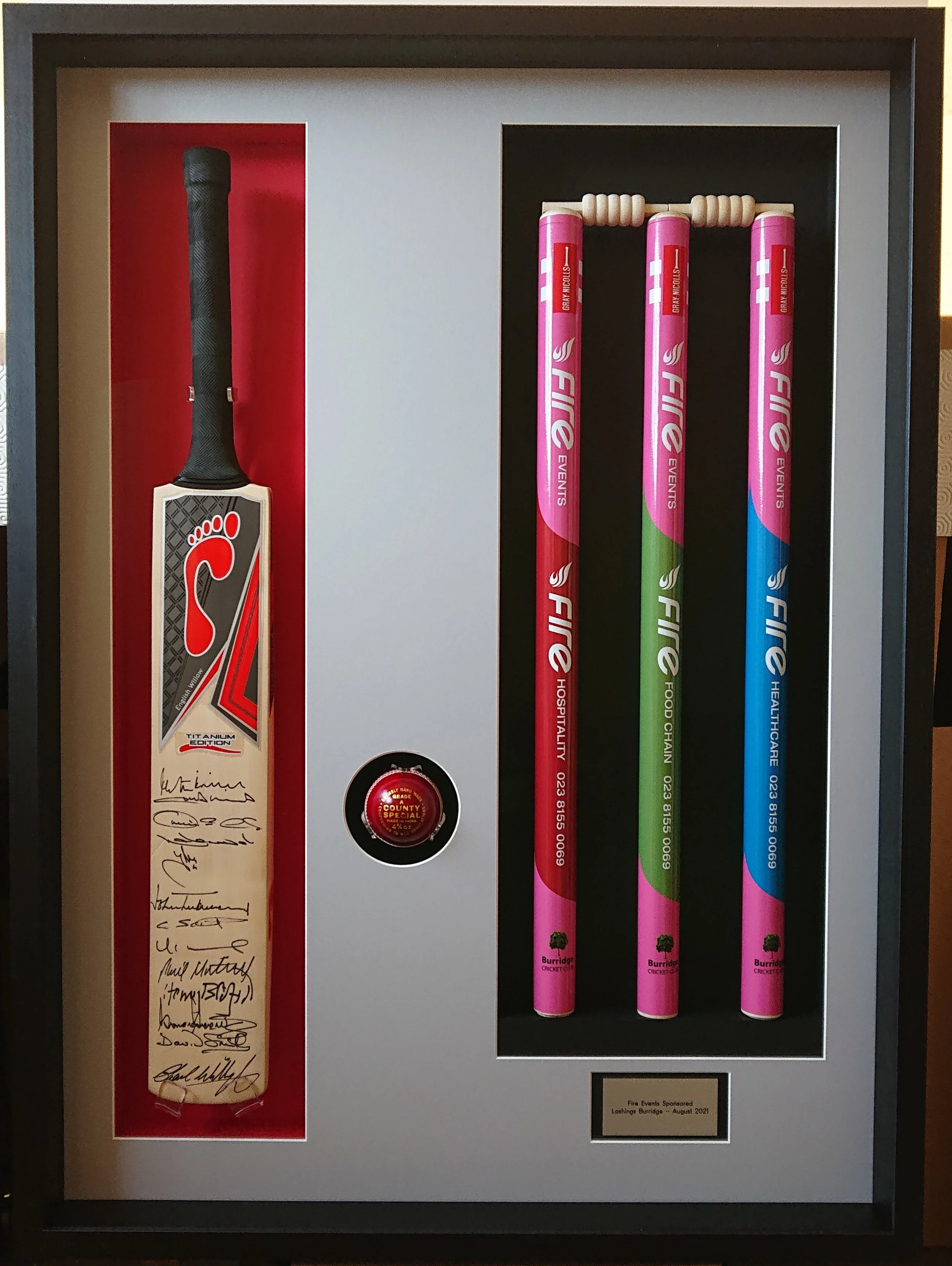 Framed display of signed cricket memorabilia, including a black cricket bat, three pink cricket stumps with sponsorship logos, a red badge, and a red and beige ribbon with signatures.