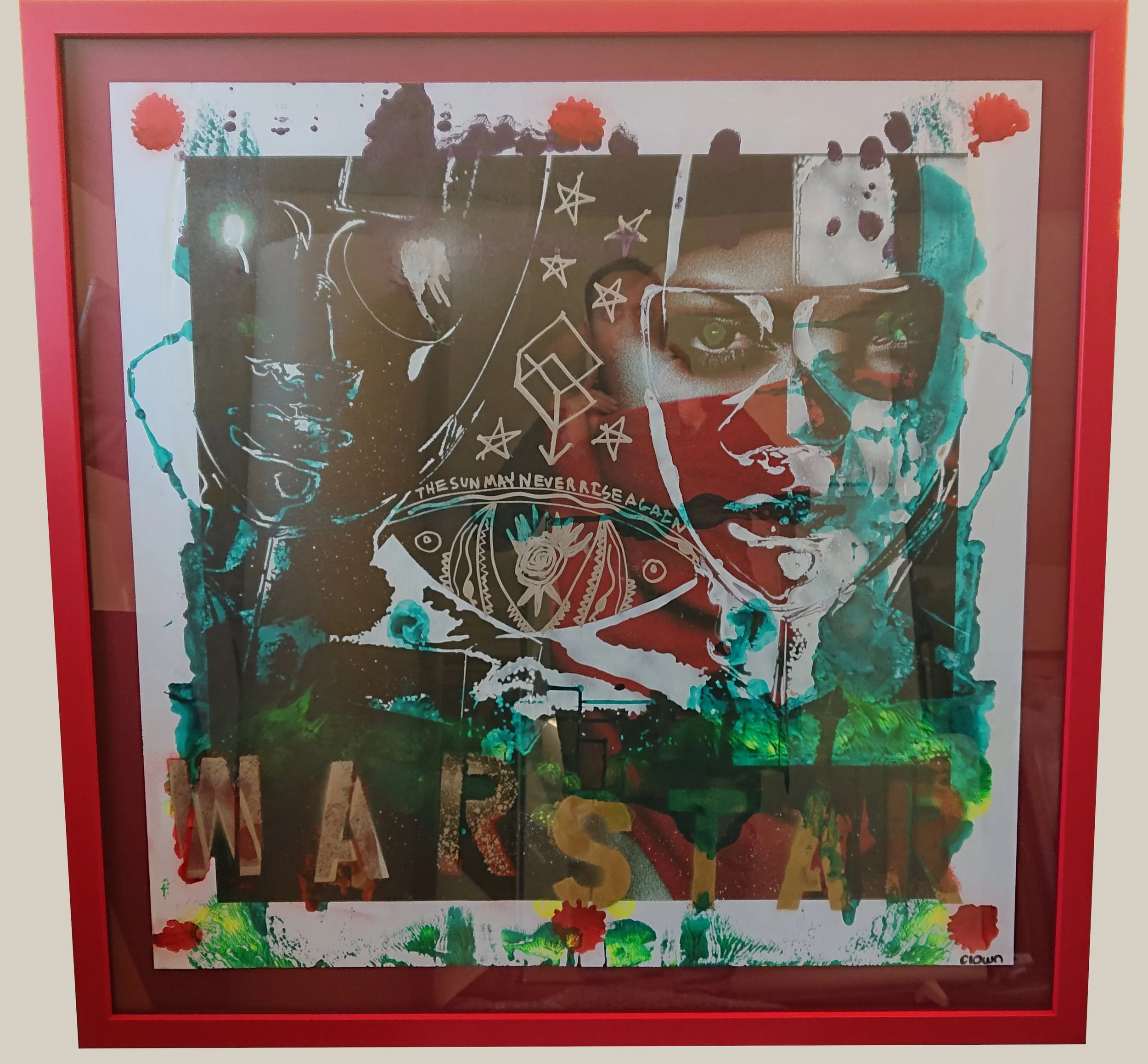 A framed abstract painting with vivid colors including reds, greens, blacks, and browns, featuring a mix of shapes, lines, and splatters. The painting includes outlined stars, hearts, and text that reads "The sun may never rise again." The frame is red, set against a neutral background.