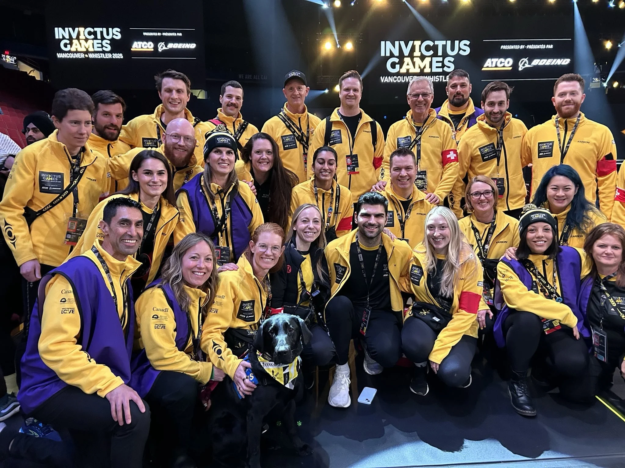 The Medical Team at the Invictus Games in Vancouver, Whistler 2025, wearing yellow and purple jackets, some with medals, posing together on stage with large screens displaying Invictus Games logo and sponsors behind them.