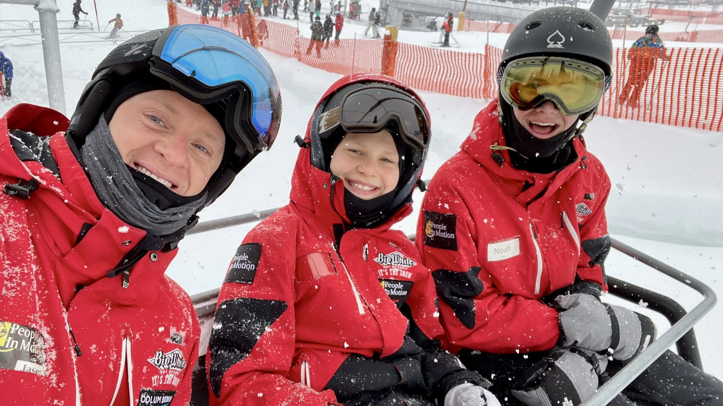 Dr. Jason with 2 young  skiers in Powderhounds red jackets and ski helmets sitting together on a ski lift, smiling, with a snowy Big White ski resort, ski slopes, and other skiers in the background.