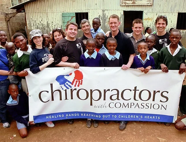 Group of Chiropractors, adults and children holding a banner that reads 'chiropractors with compassion' outside a rustic building in the area of Mathare in  Nairobi Kenya, smiling for the camera.