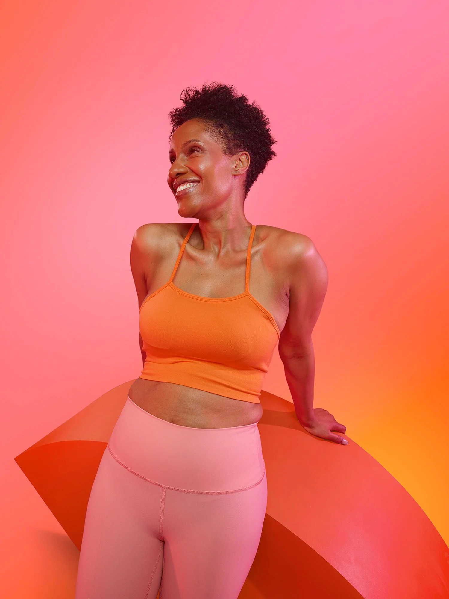 A woman with short curly hair smiling and looking to her left, wearing an orange sports top and light pink leggings, standing against a pink and orange gradient background.