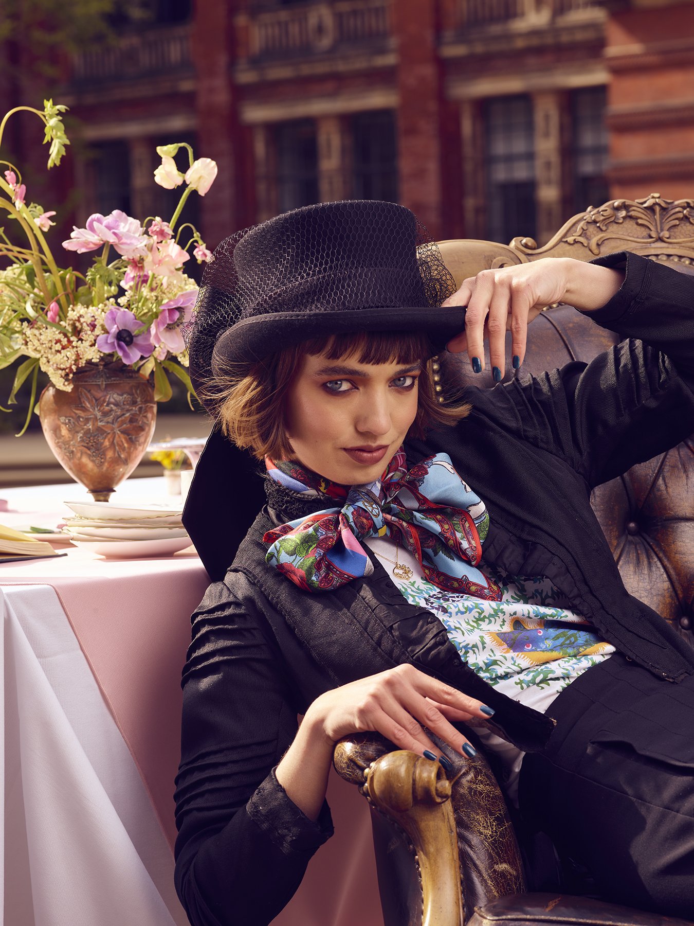 Woman in vintage attire with top hat and colorful scarf, sitting on a vintage chair beside a table with a vase of flowers.