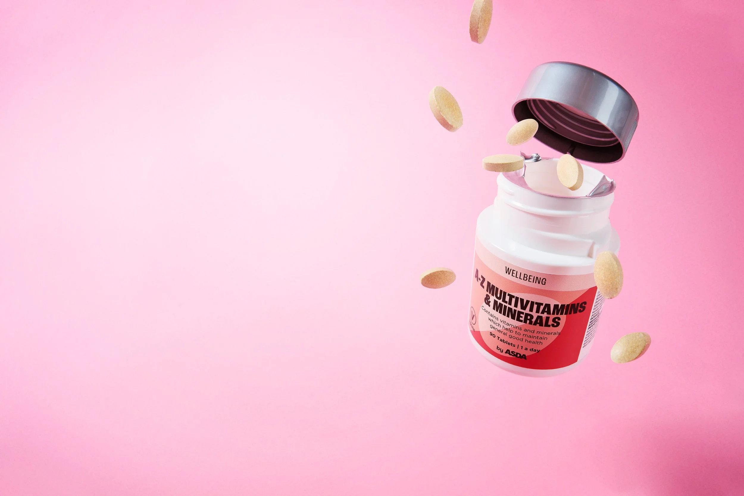 Open bottle of multivitamin tablets on pink background with tablets falling out.