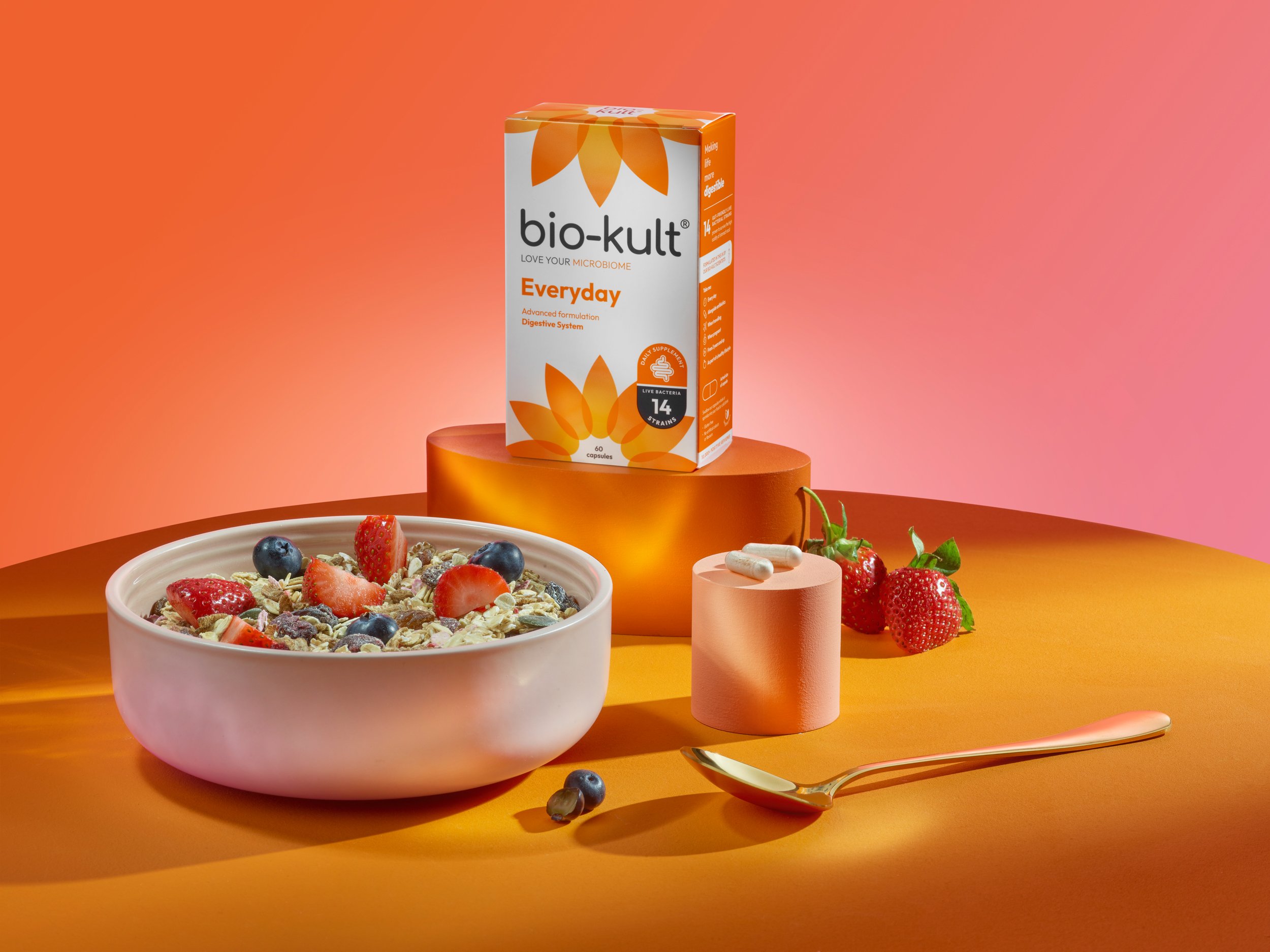 A bowl of cereal with strawberries and blueberries, strawberries on the table, a box of Bio-Kult Everyday probiotic capsules on an orange pedestal, a pink cylindrical container with tablets, a gold spoon on an orange table, and a pink background.