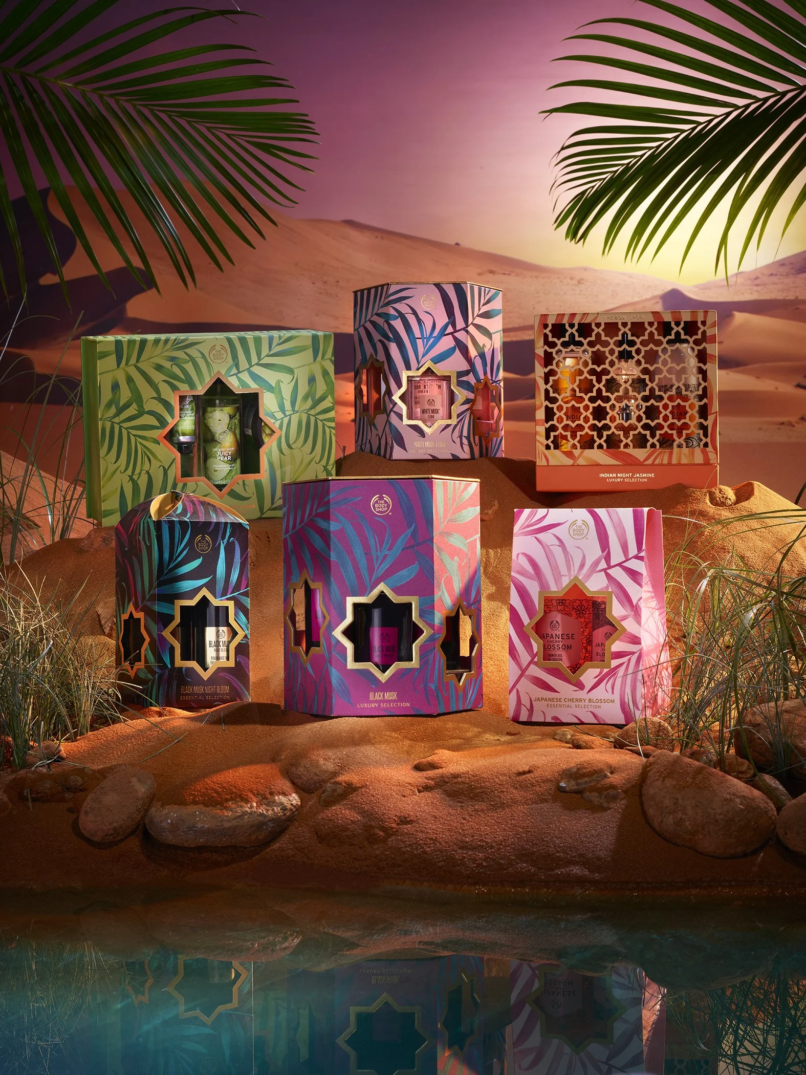 Colorful boxed set of scented products displayed on desert terrain with palm fronds and a sunset in the background.