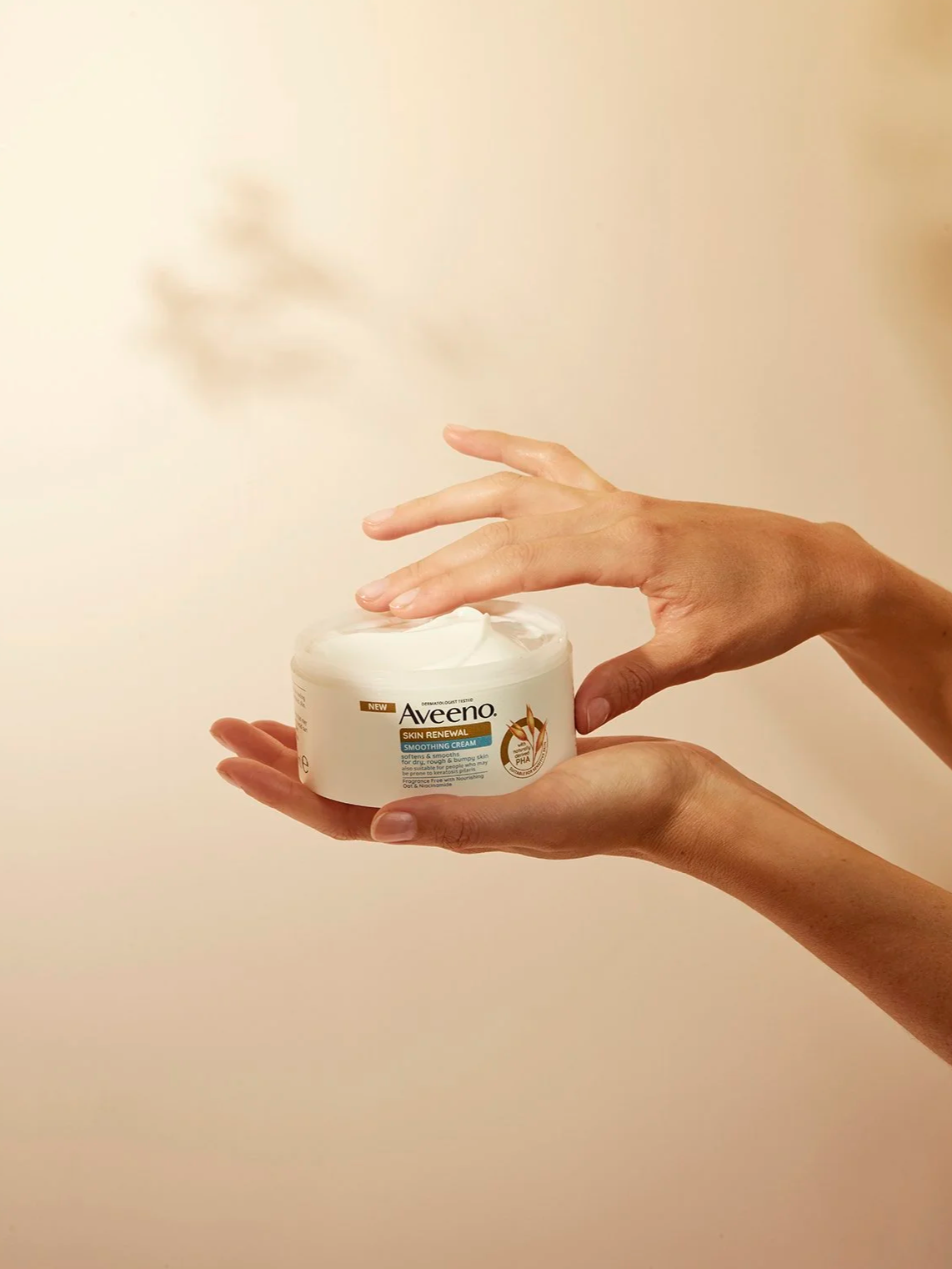 Hands holding a container of Aveeno Skin Renewal Smoothing Cream with a plain beige background.