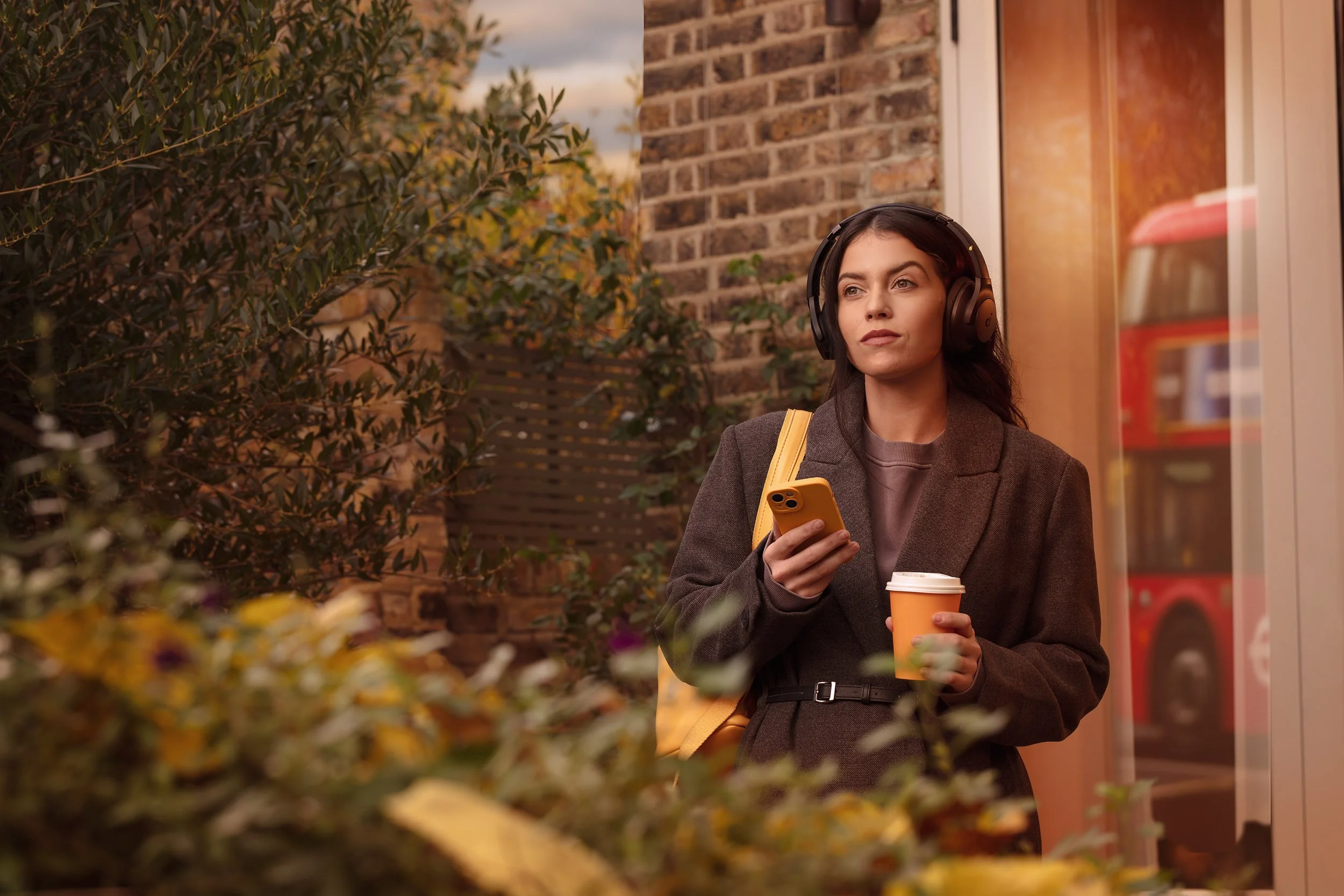 A woman wearing a coat and headphones, holding a coffee cup and looking off to the side outside a store or café in the fall, with brick walls and trees with autumn leaves in the background.