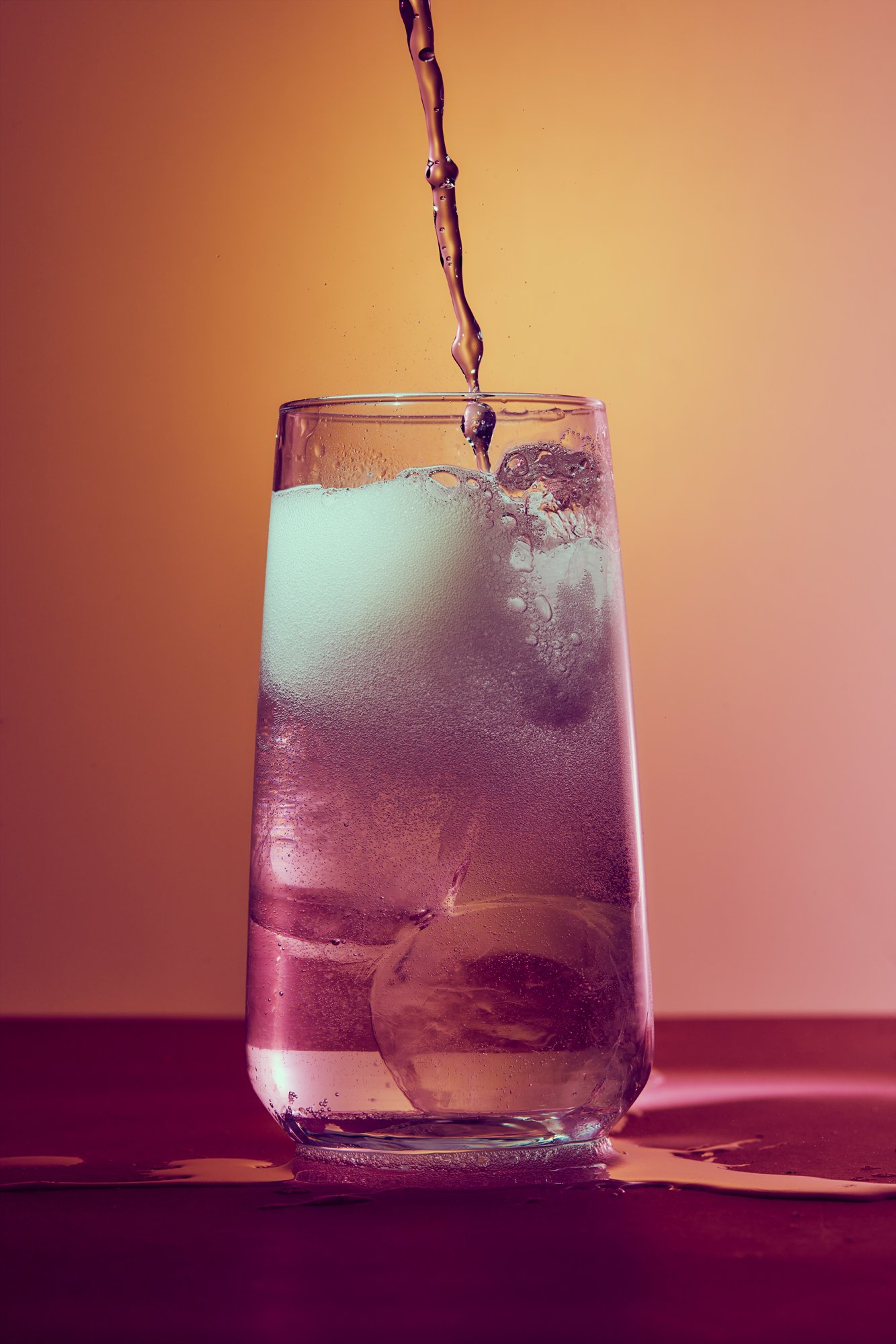 A glass of water with ice and bubbles being poured into it on a purple surface with a pink and orange gradient background.