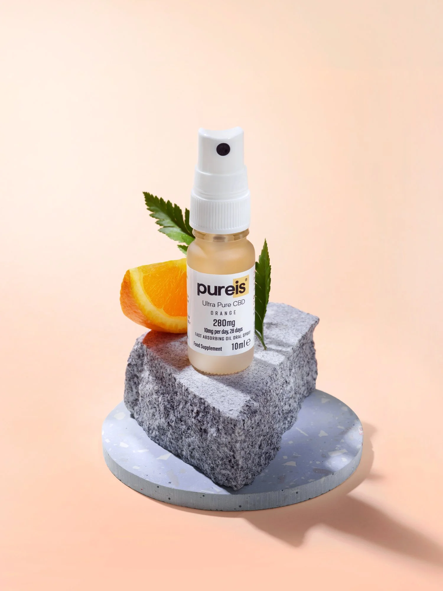 A small bottle of Pureis ultra pure CBD spray with orange flavor, placed on a gray stone block with a slice of orange and green leaves for decoration, on a round gray base with a plain peach-colored background.