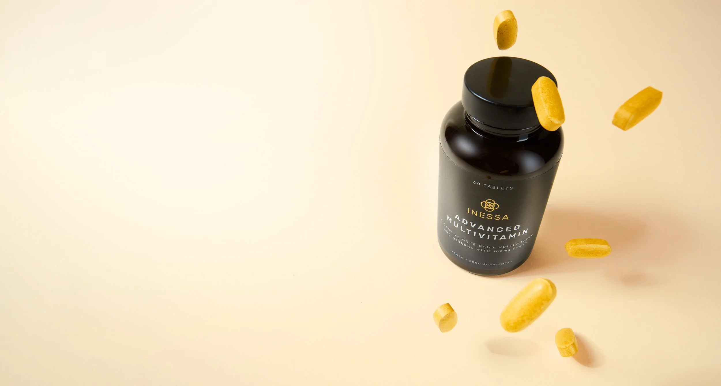 A black bottle labeled 'Inessa Advanced Multivitamin' with yellow capsules floating around it on a beige background.
