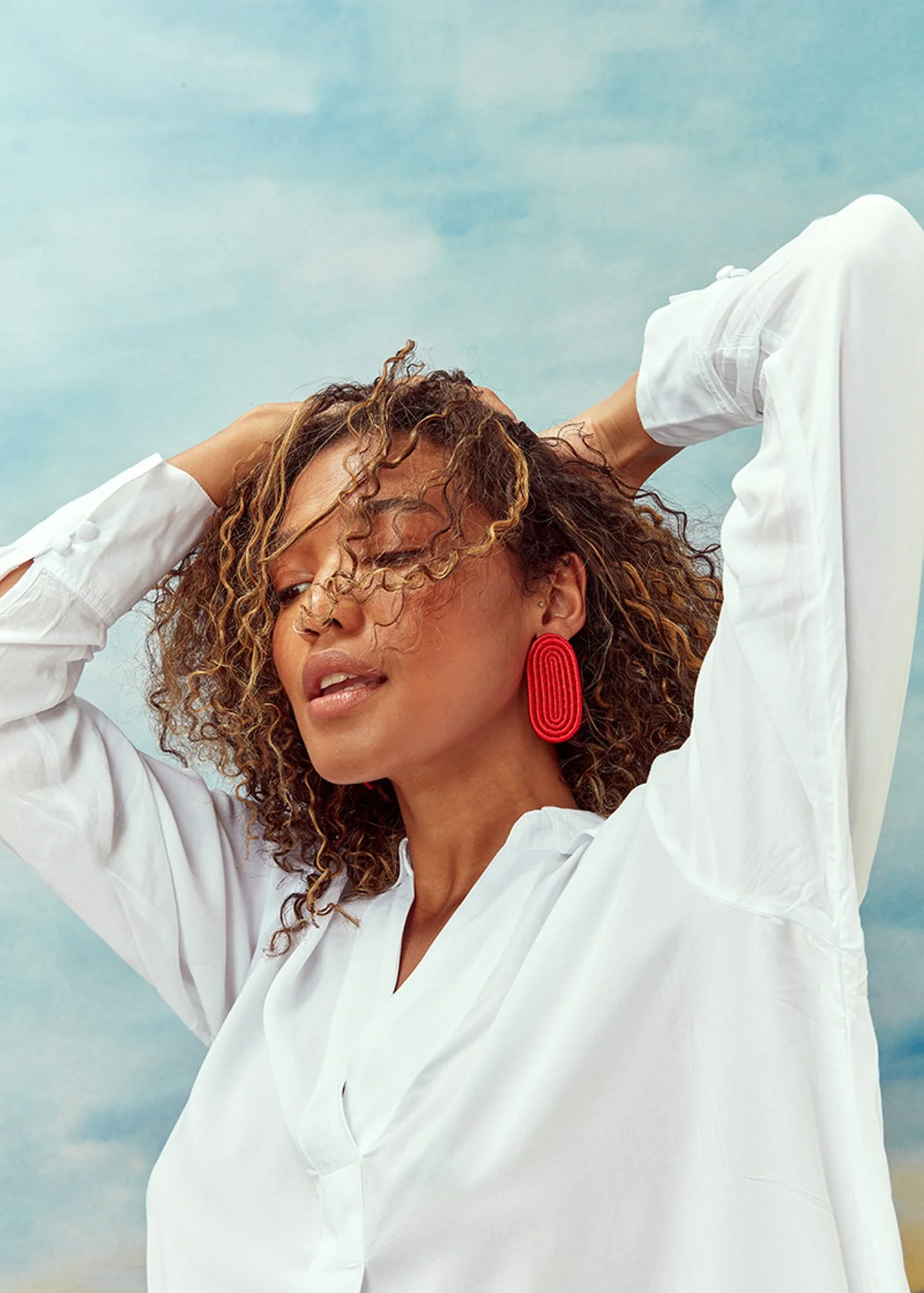 A woman with curly hair wearing a white blouse and large red earrings, posing outdoors with her eyes closed and hands behind her head under a cloudy sky.