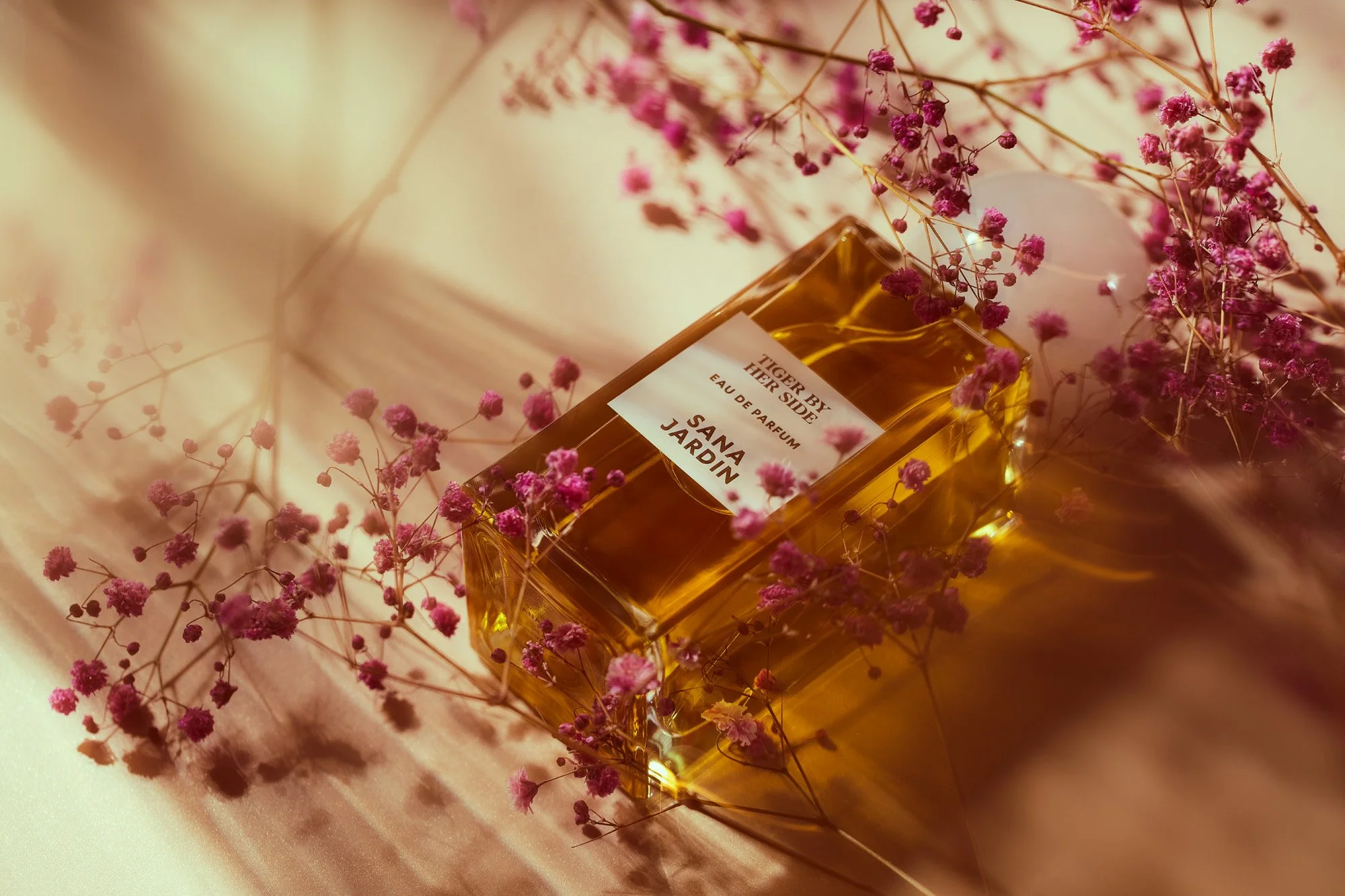 A perfume bottle labeled 'Tiger By The Side Eau de Parfum Sanja Jardin' surrounded by pink baby's breath flowers, with a soft warm background.