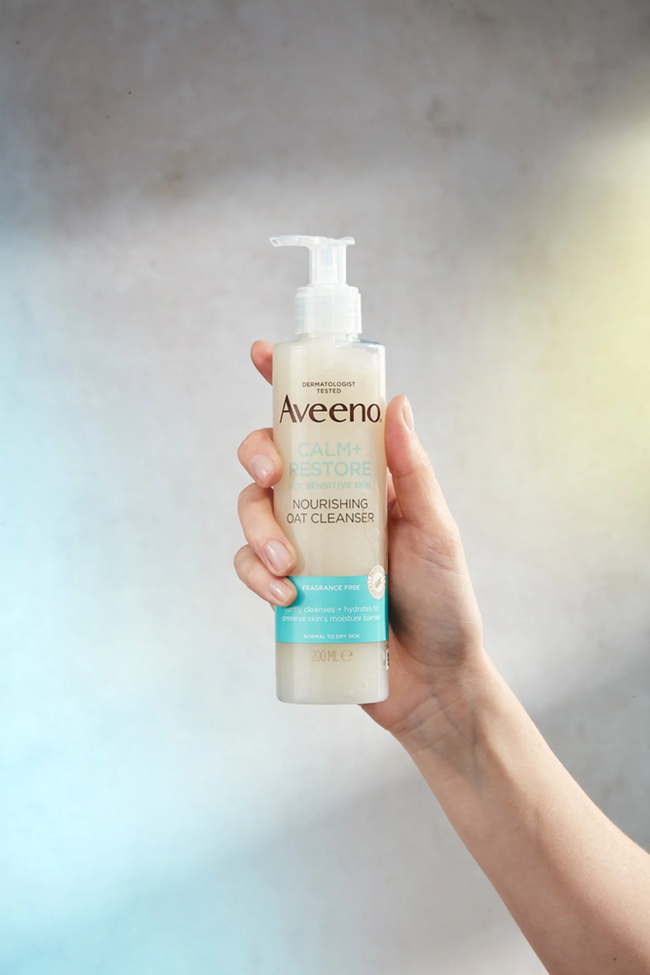Hand holding a bottle of Aveeno Calm + Restore Nourishing Oat Cleanser for sensitive skin.