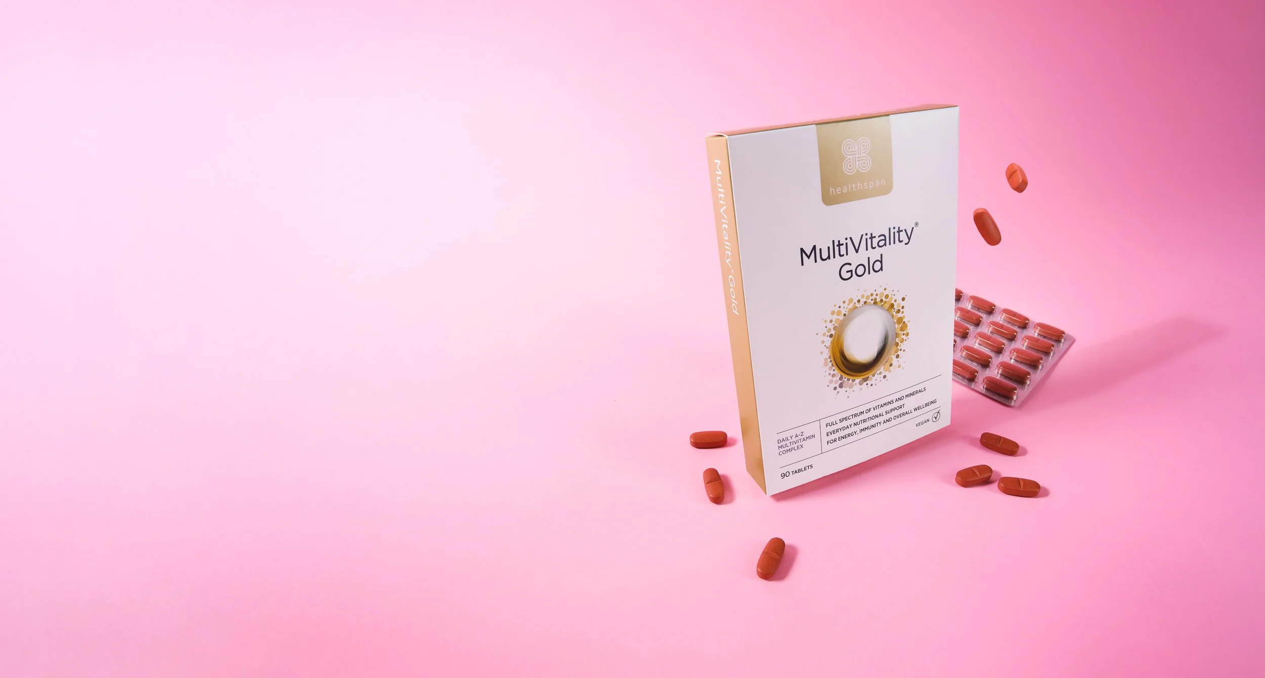 A pink background with a box labeled 'MultiVitality Gold' supplement, surrounded by scattered reddish-brown tablets and a blister pack containing more tablets.