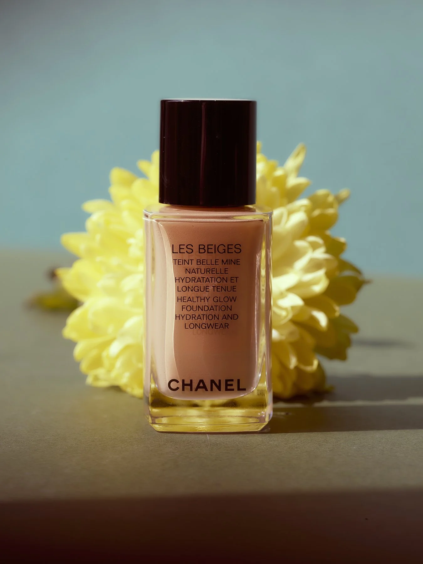 A bottle of Chanel Les Beiges foundation with a yellow flower behind it, set against a blurred background.