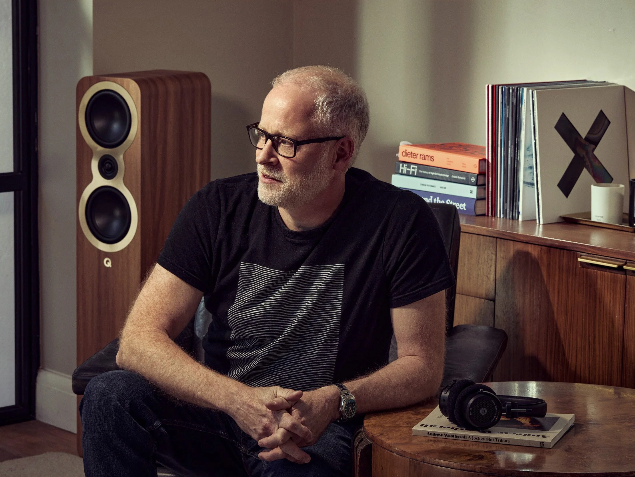 A middle-aged man with glasses, a beard, and light-colored hair, sitting at a wooden table in a room with books and a large speaker in the background. He is wearing a black t-shirt and a watch, with headphones and a book on the table in front of him.