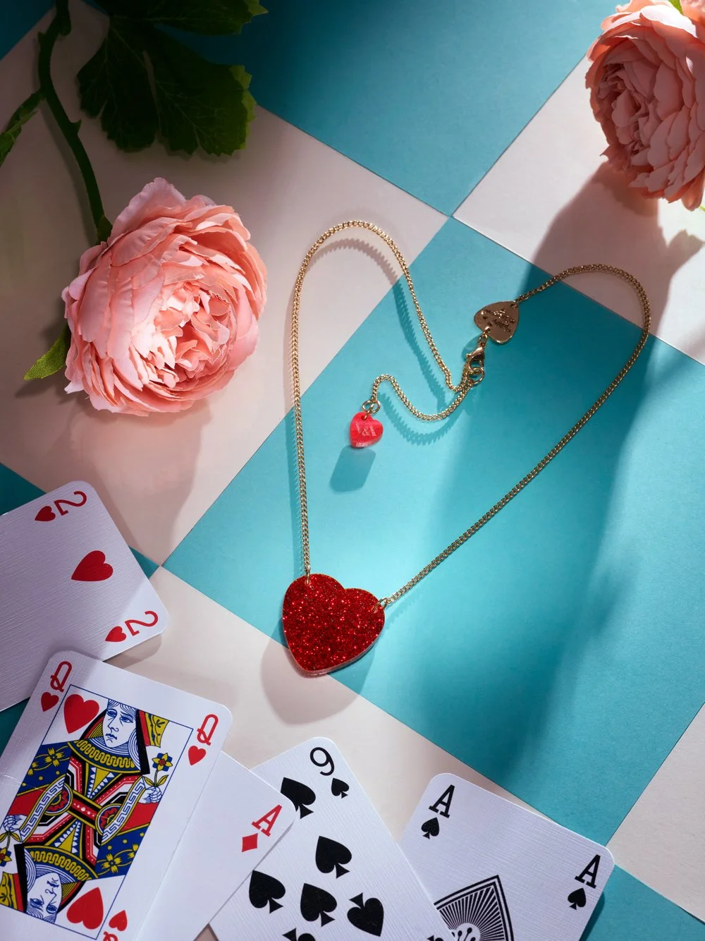 A flat lay image featuring a red glittery heart-shaped pendant on a gold chain, surrounded by playing cards and pink peonies, set against a checkered blue and white background.