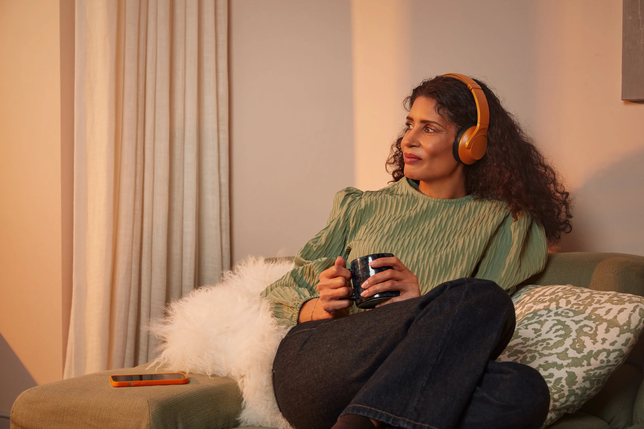 A woman with curly dark hair wearing a green striped top and headphones sitting on a sofa, holding a ceramic mug, in a cozy room with beige curtains and a plush white throw pillow.