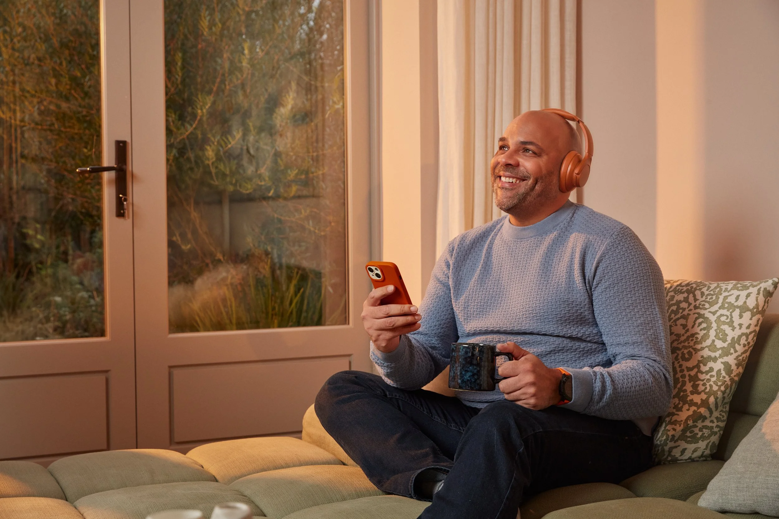 A man sitting on a beige couch during sunset, wearing headphones, holding a coffee mug, smiling at his phone with a window in the background showing trees outside.