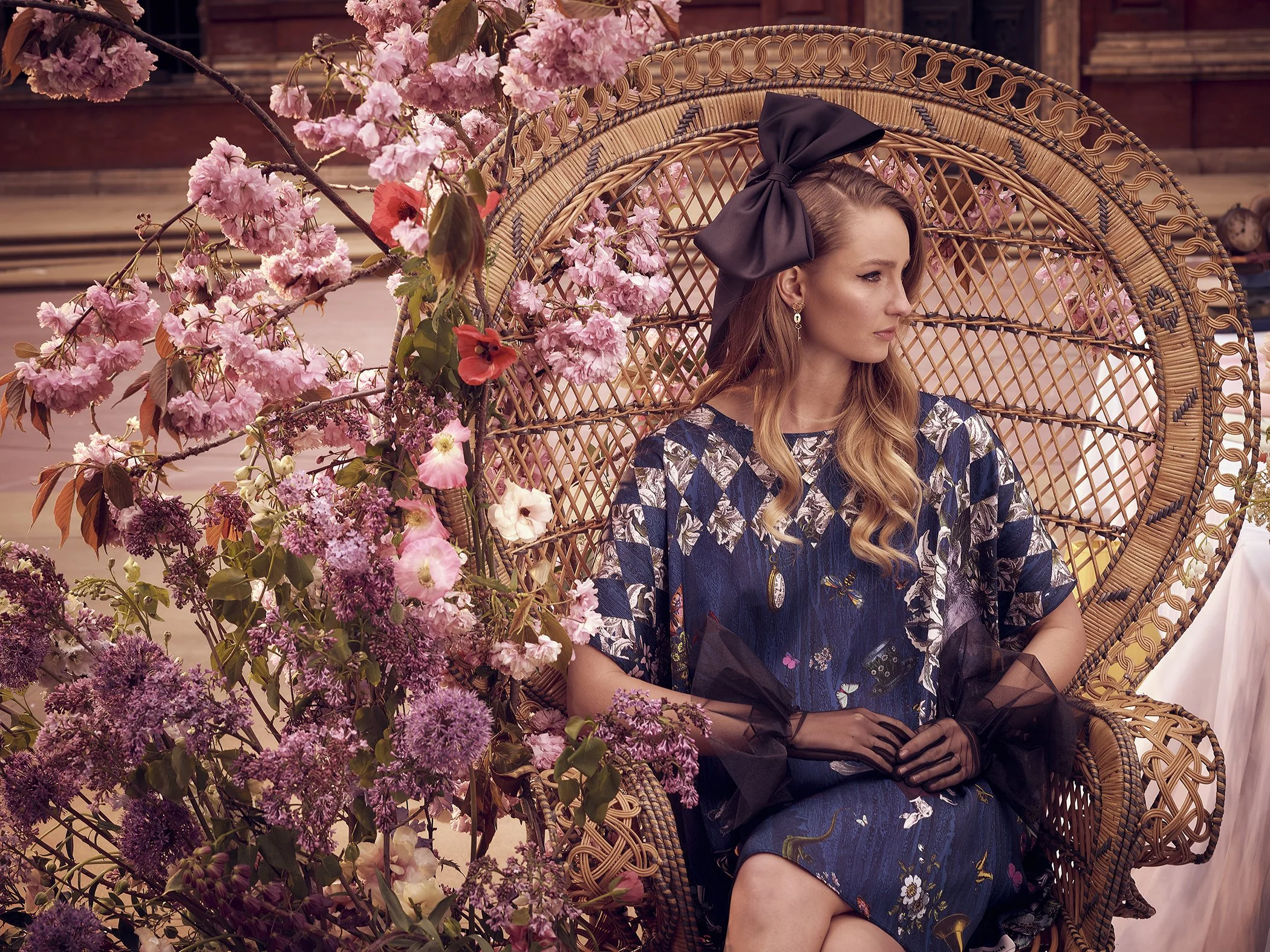 Woman in floral dress and large bow headband seated in wicker chair, surrounded by pink flowering branches.