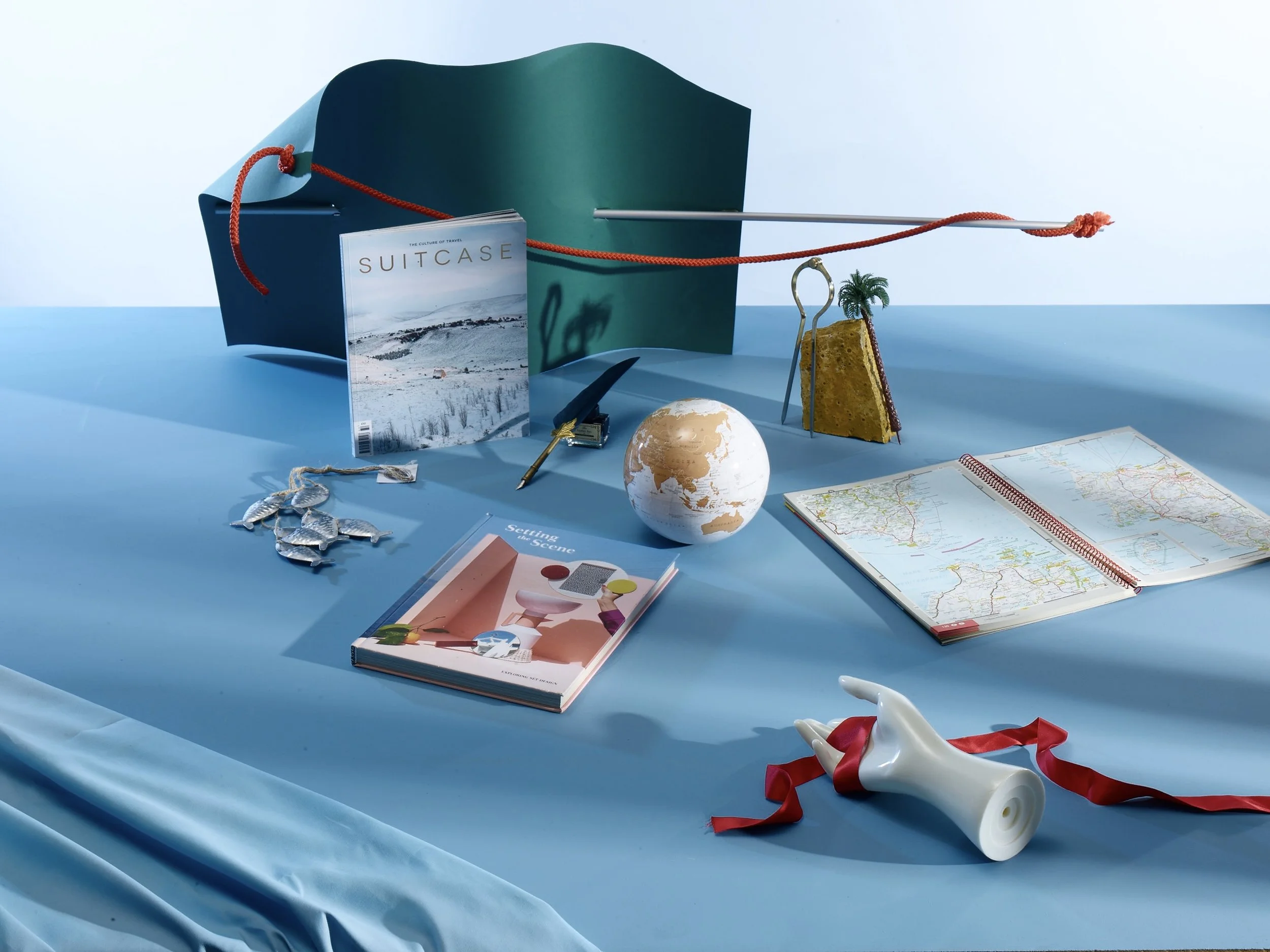 Travel-themed objects arranged on a blue surface, including a suitcase, a magazine titled "Suitcase," a map, a globe, a book, a necklace with fish charms, a small sculpture of a human hand holding a megaphone with a red ribbon, a slice of sponge cake