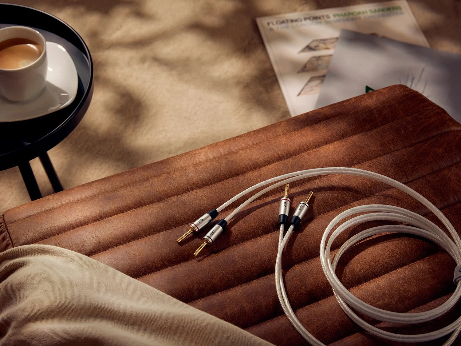 A pair of high-end audio cables with gold-plated connectors placed on a brown leather armrest, with a small white cup of espresso on a black tray in the background and a pile of magazines and papers nearby.