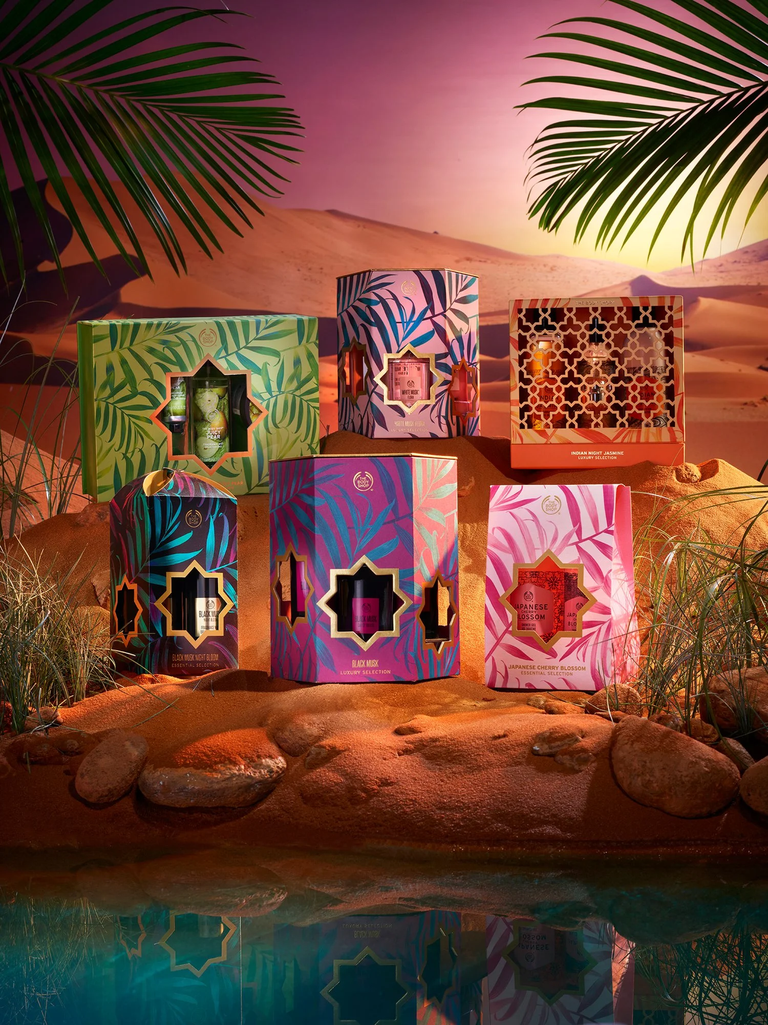 Colorful decorative boxes with tropical leaf patterns arranged on desert rocks, with palm leaves and desert dunes in the background during sunset.