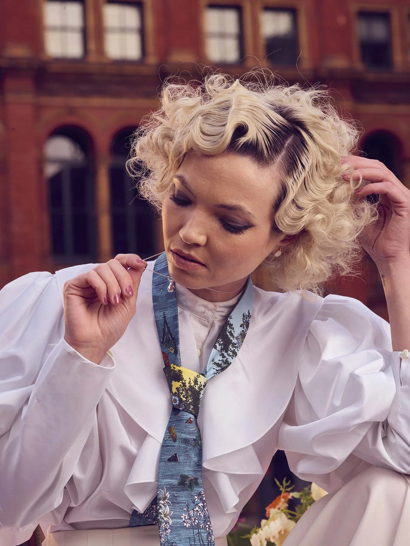 Model with curly blonde hair in a white blouse and patterned tie, posing outdoors in front of brick building.