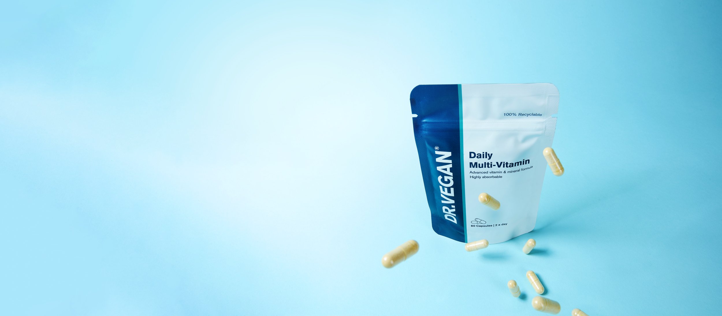 A blue and white pouch labeled 'Dr. VEGAN Daily Multi-Vitamin' standing on a blue background with several yellow capsules scattered in front.
