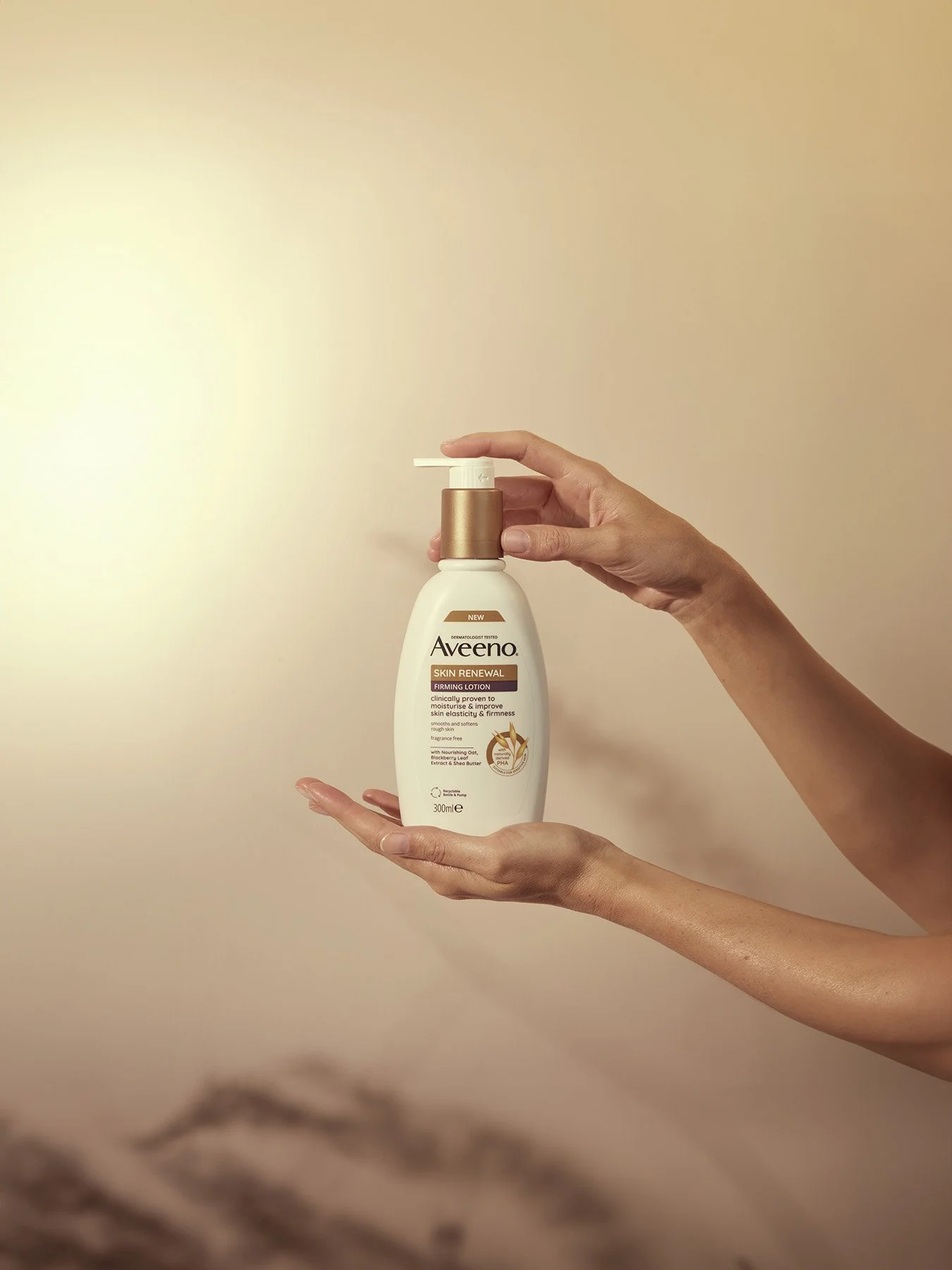 A person holding a bottle of Aveeno Skin Renewal Firming Lotion with a pump dispenser against a plain beige background.